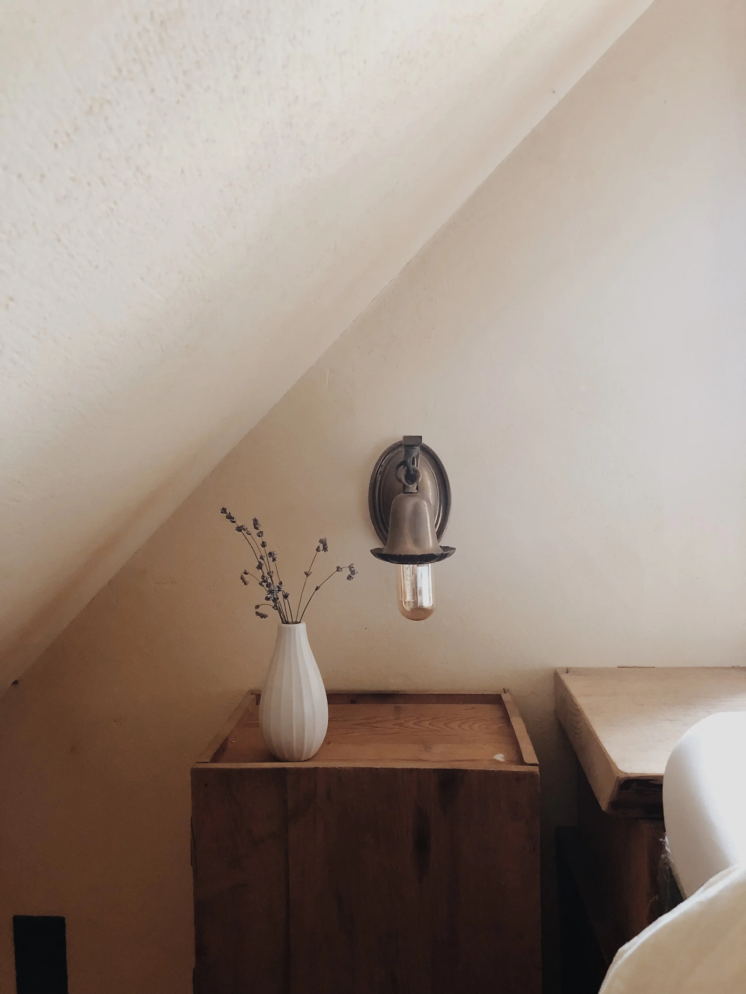 Wooden cabinet with white vase and dried lavender, wall-mounted lamp on sloped ceiling wall in a cozy room.