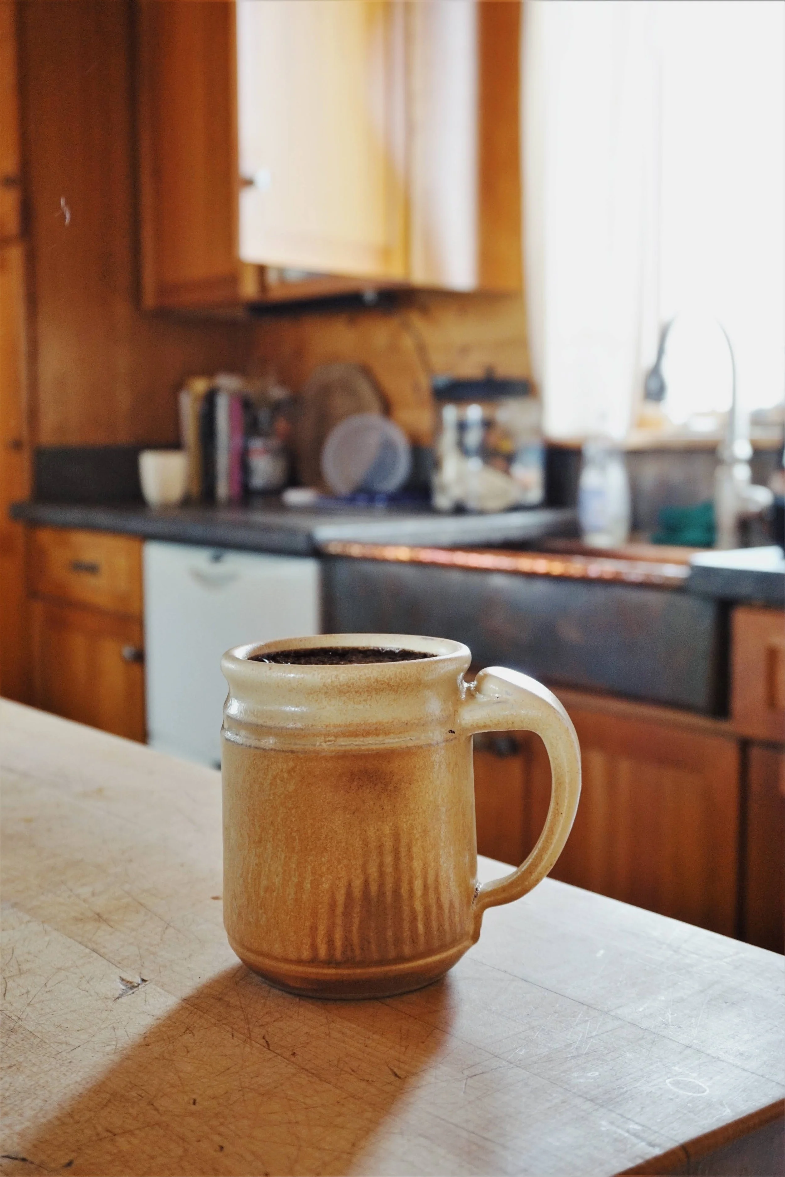 A ceramic mug filled with coffee sits on a wooden kitchen counter with a rustic kitchen in the background.