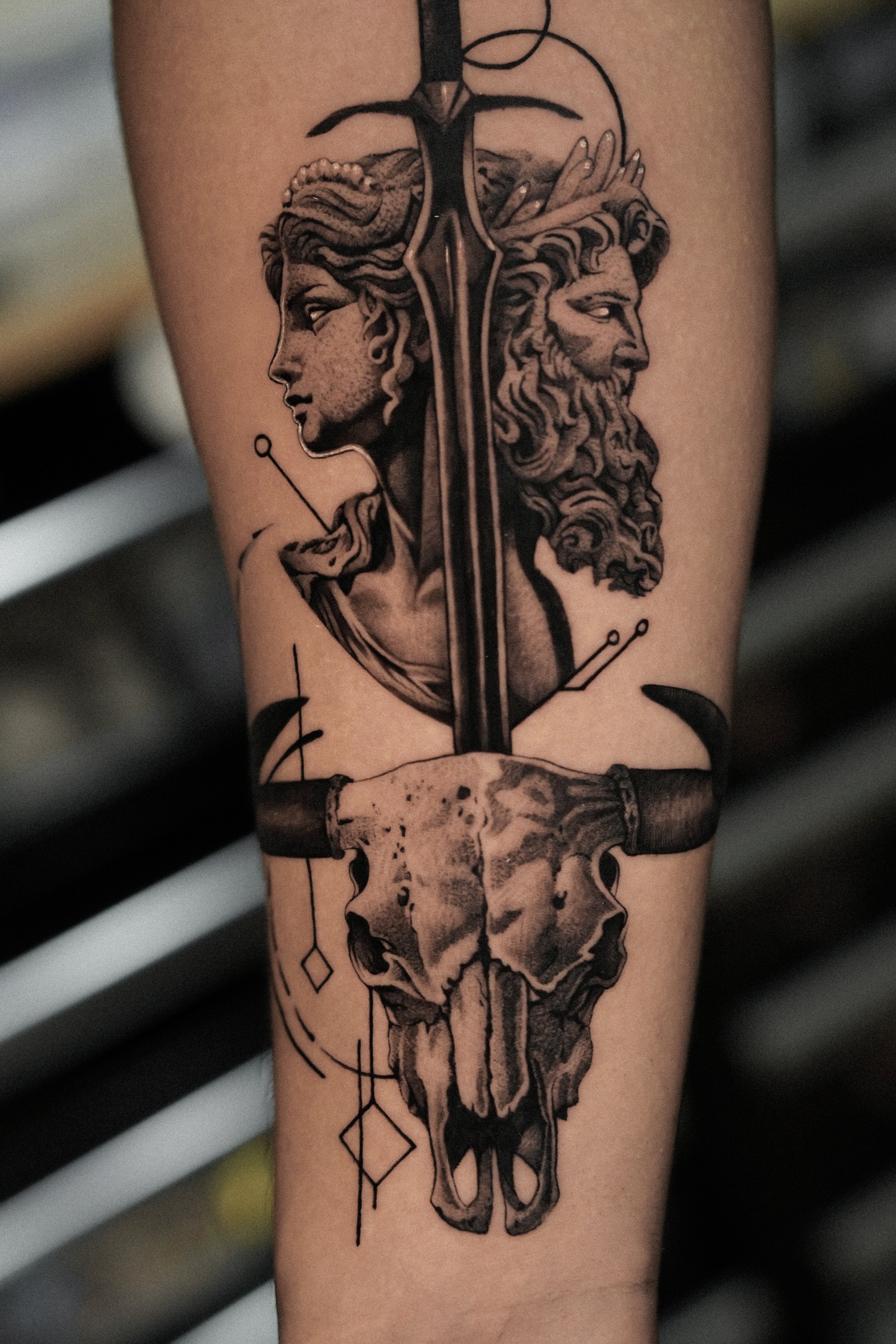 Tattoo featuring a sword between two faces, male and female, and a skull with horns at the bottom.