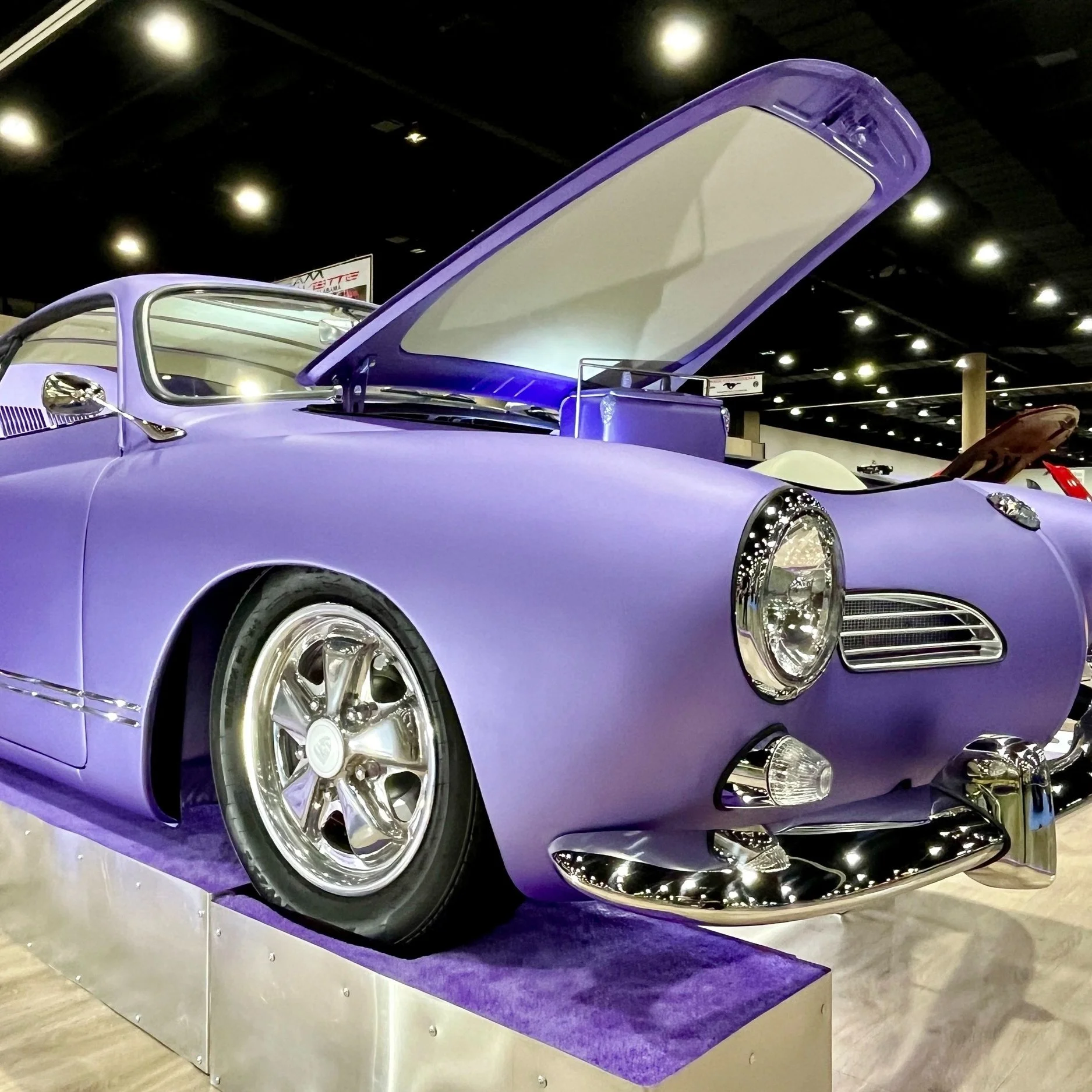 Classic purple car with hood open, showcasing chrome details and round headlights, displayed at indoor auto show with bright lighting.