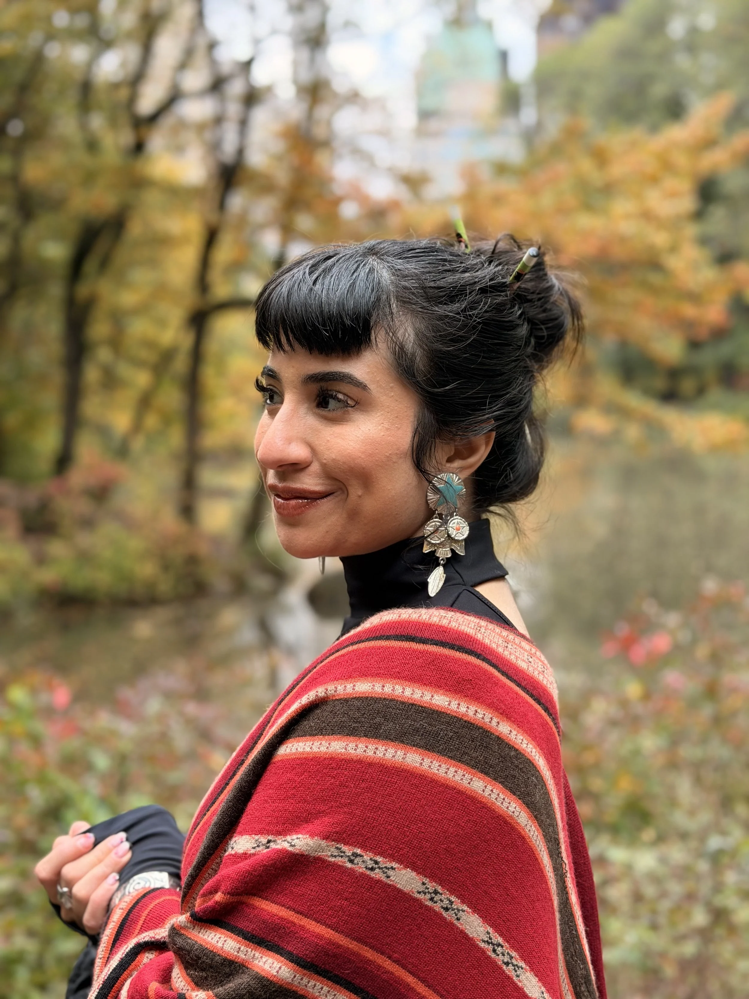A woman with black hair and bangs, wearing large turquoise and silver earrings, a black turtleneck, and a red, orange, and brown striped shawl, standing outdoors with autumn trees and a body of water in the background.