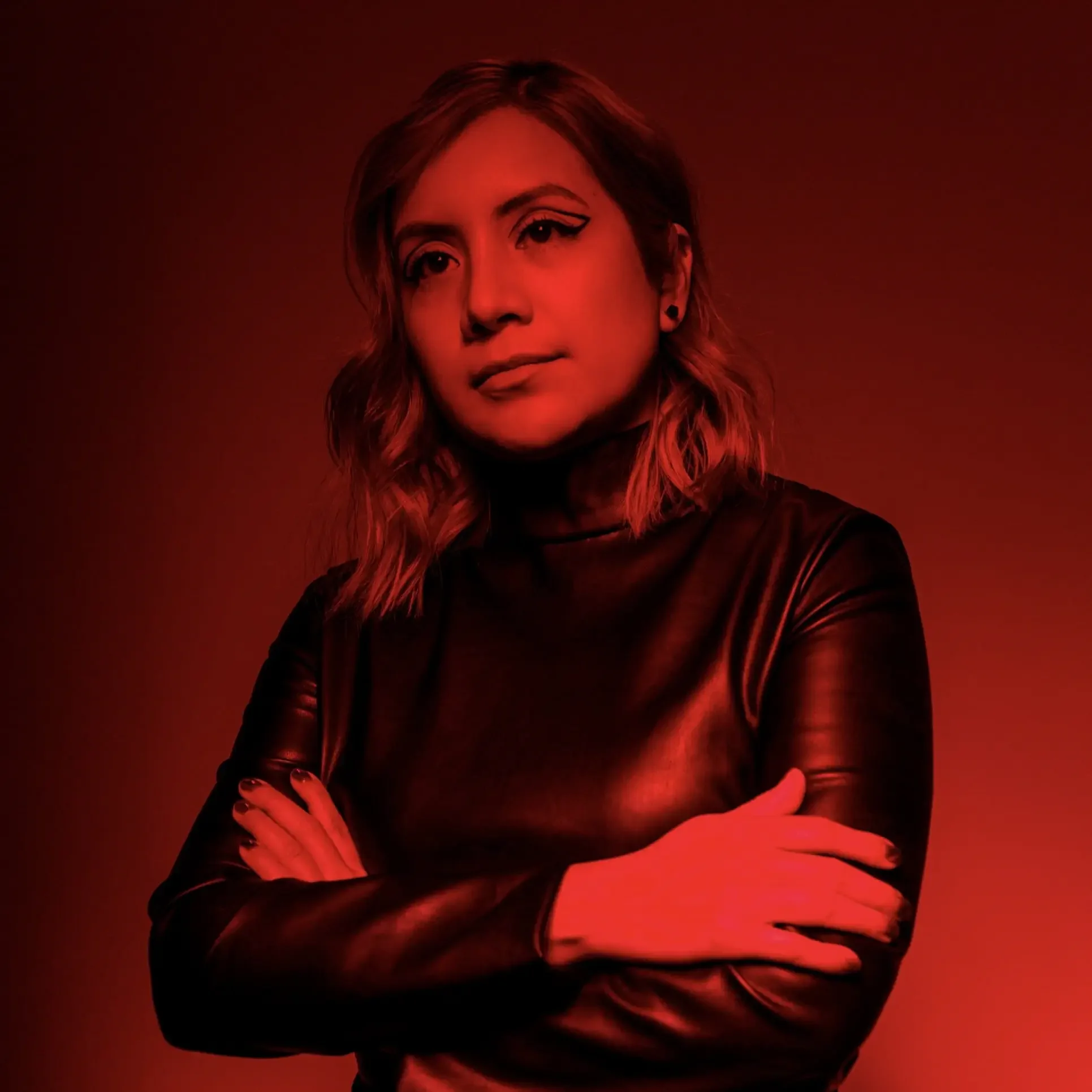 A woman with shoulder-length wavy hair, in a black leather top, looking at the camera with her arms crossed, illuminated by red lighting.