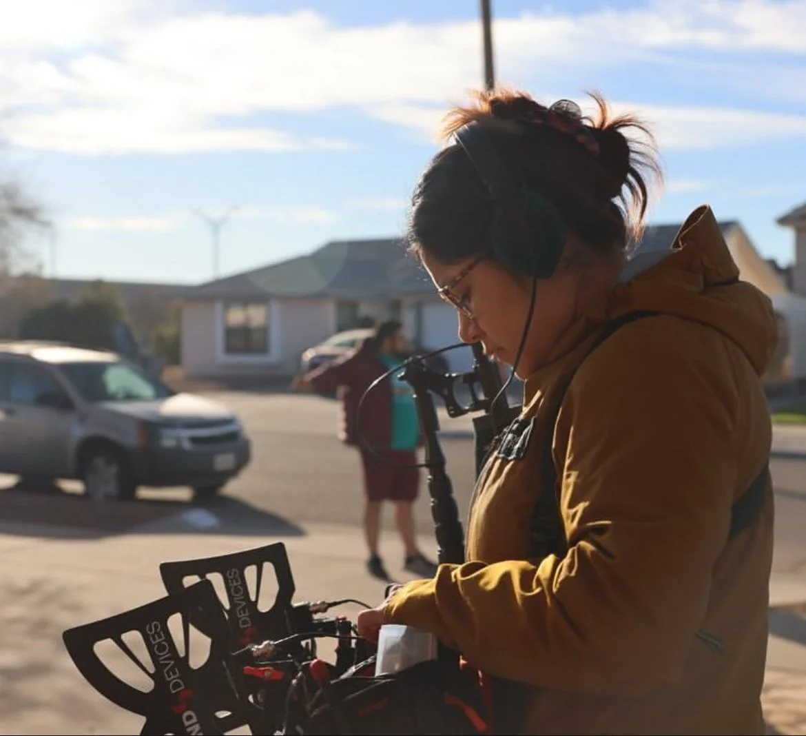 A person with glasses and headphones, wearing a brown jacket, is looking down at a bicycle in a suburban neighborhood during daytime.