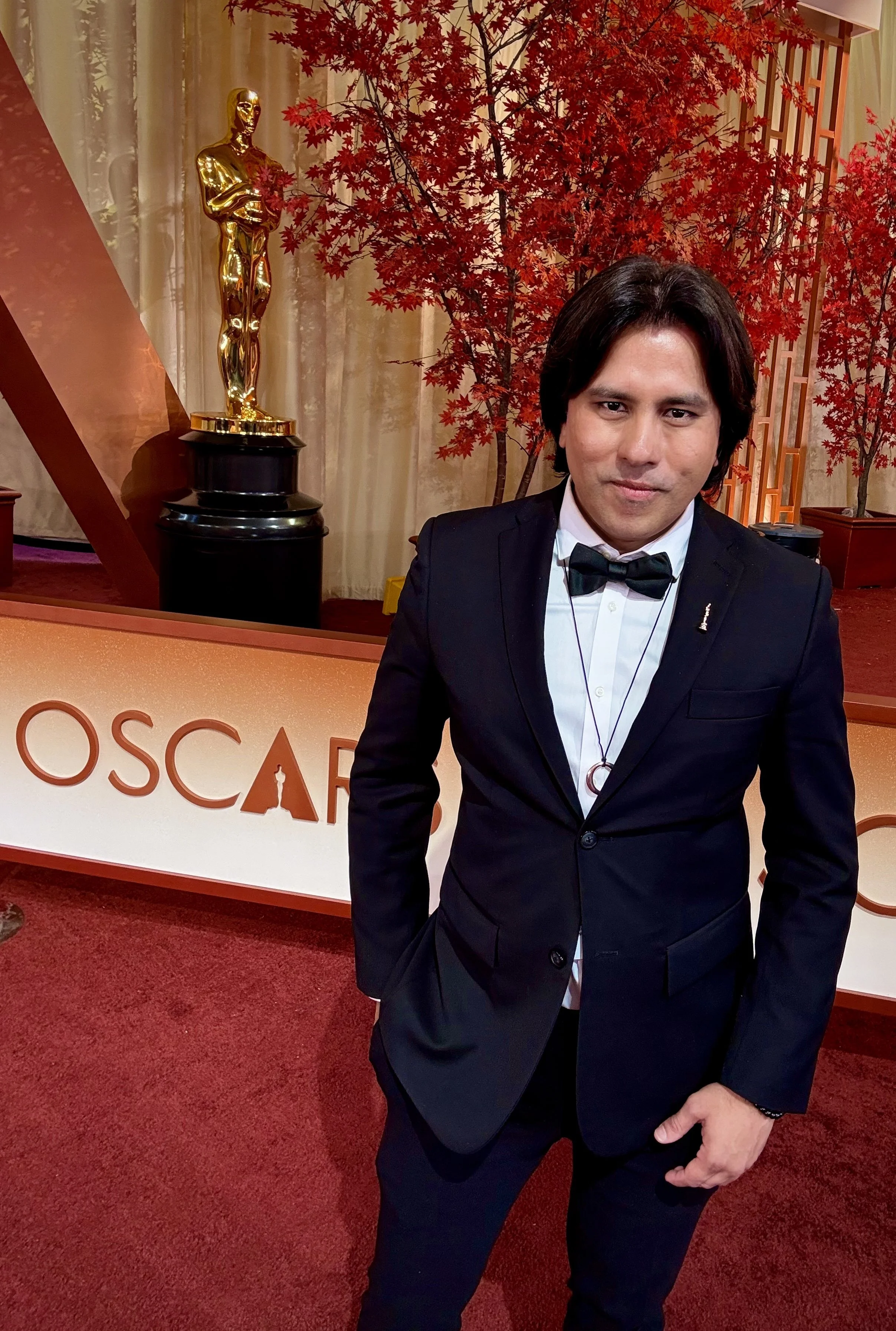 A man in a tuxedo with a bow tie at an awards event, standing in front of a sign with the Oscar logo and a decorative background with a red tree sculpture and an Oscar statue.