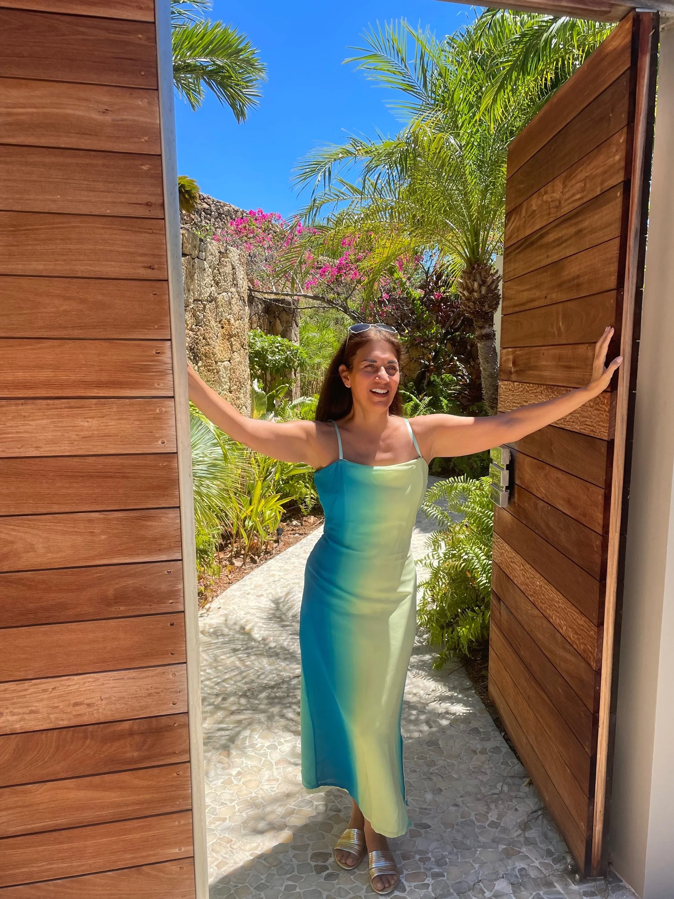 Maggie Franco standing outdoors opening large wooden doors, smiling in a flowing blue-green dress, surrounded by tropical plants and sunlight.