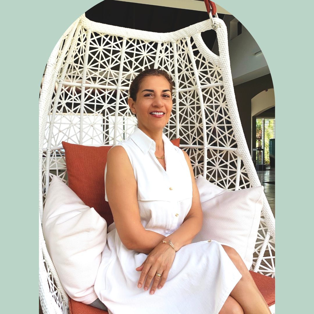 Woman sitting on a white woven hanging chair with white and rust-colored cushions, smiling, wearing a sleeveless white dress.