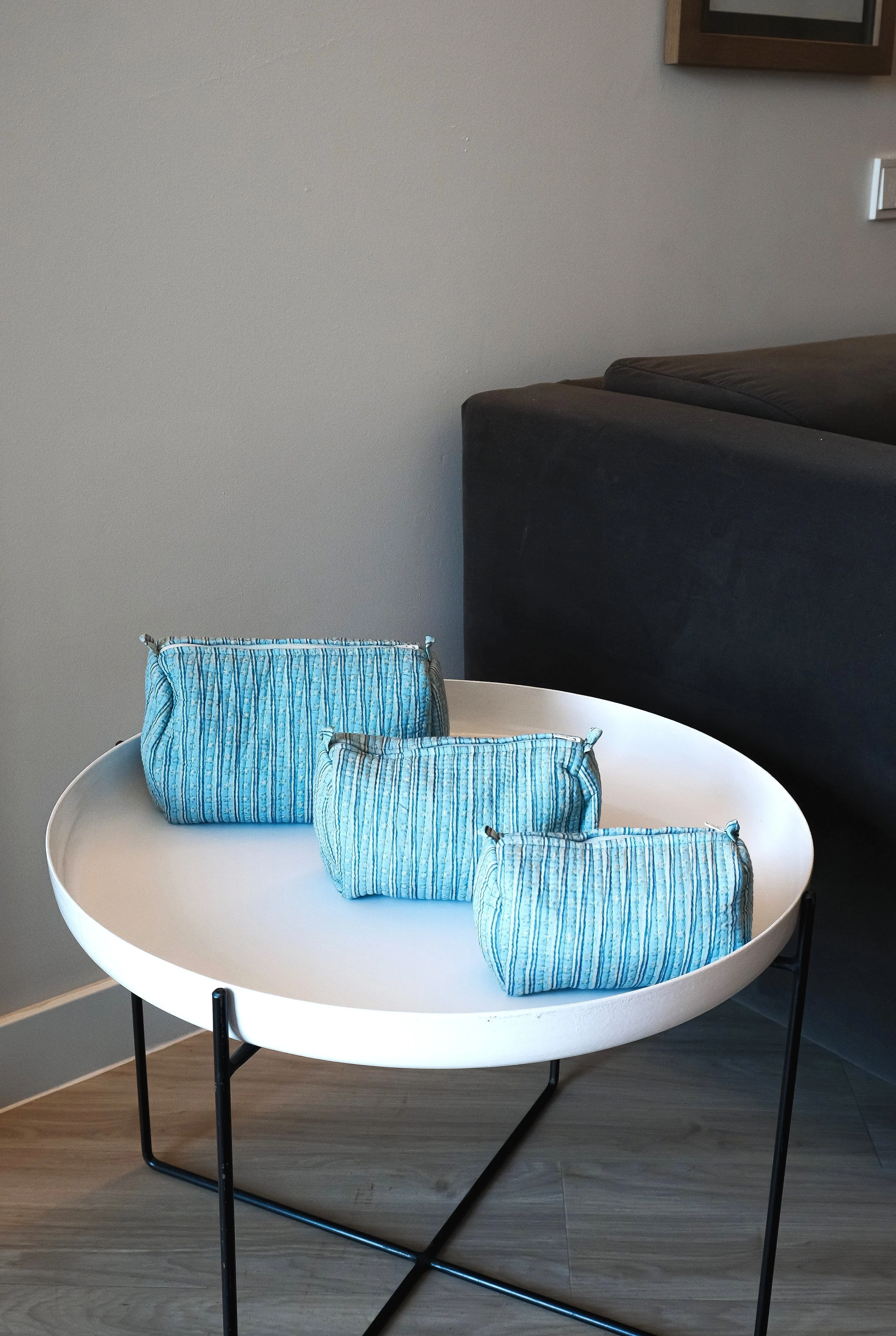 Tres bolsas de tela azul en una mesa blanca redonda con patas negras, junto a un sofá gris oscuro.