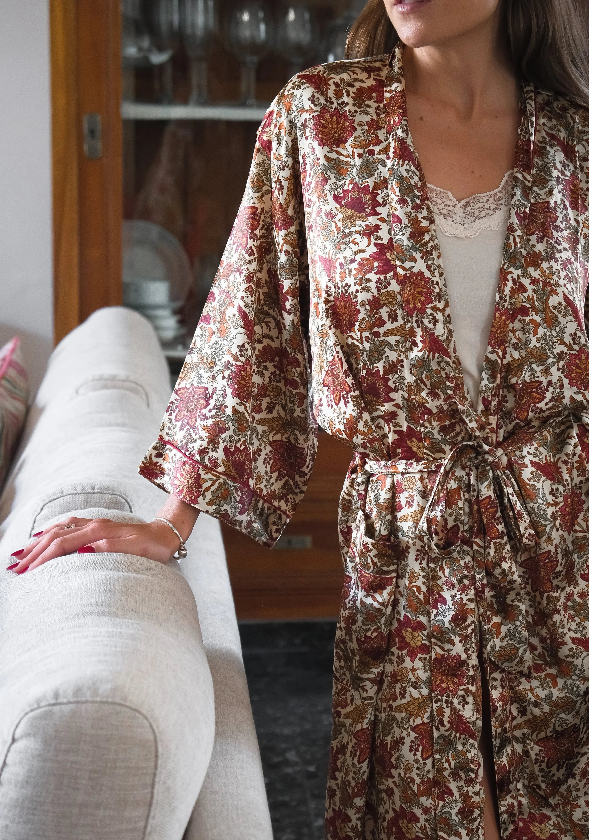 Mujer con bata floral colorida, apoyada en un sofá beige, en una sala con estantería de fondo.