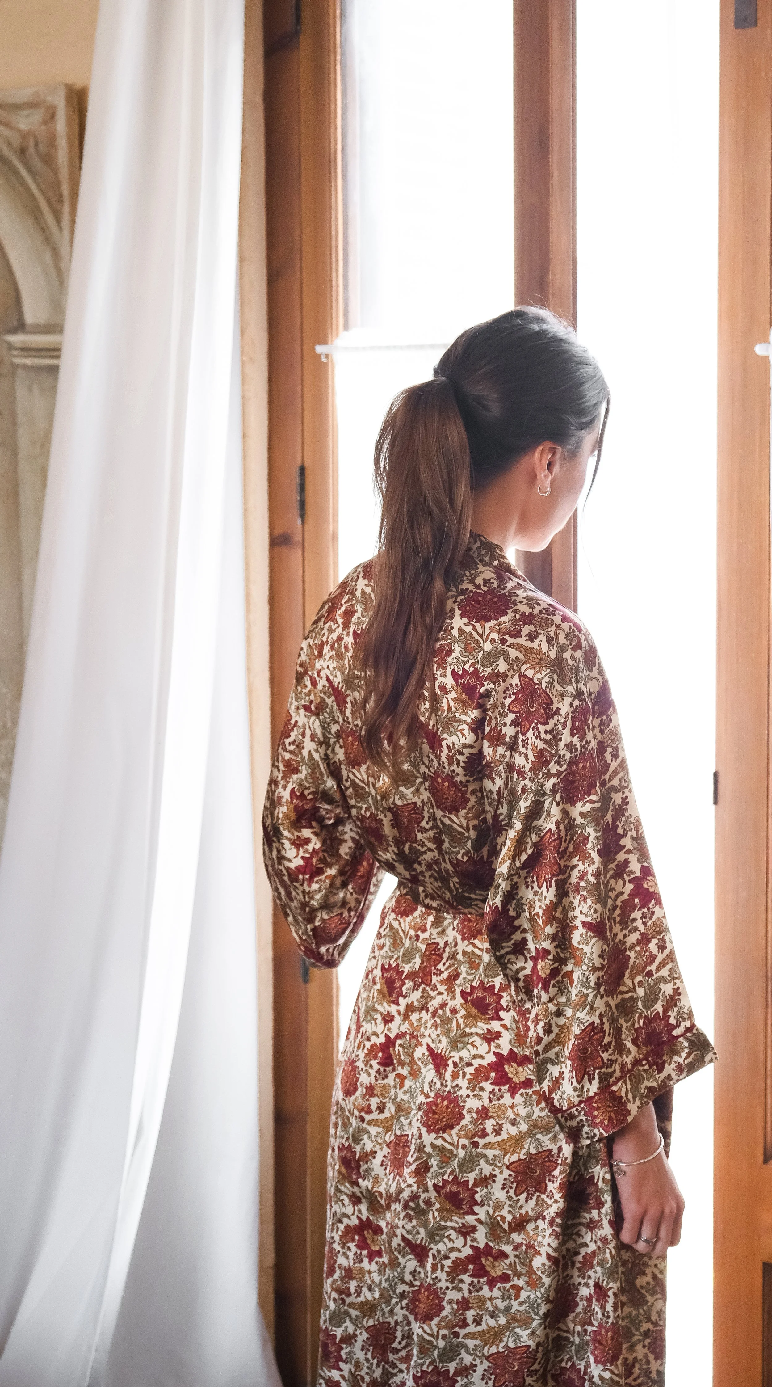 Mujer de espaldas con bata floral mirando por la ventana.