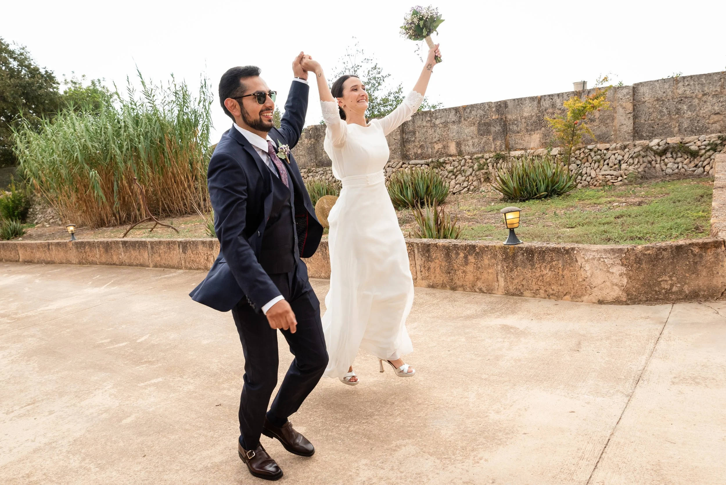 Pareja recién casada caminando juntos, ella con vestido blanco y ramo de flores, él con traje oscuro y gafas de sol, en un entorno al aire libre con vegetación y un muro de piedra.