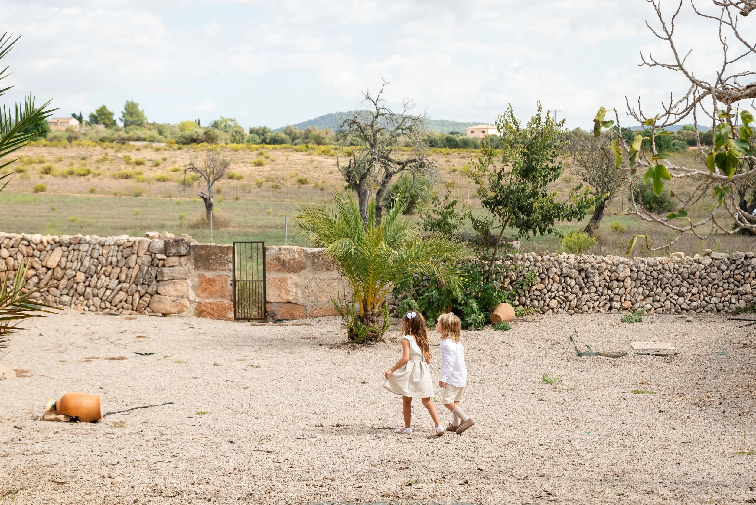 Dos niñas caminando en un patio de piedra con un muro de piedras al fondo y un paisaje rural.