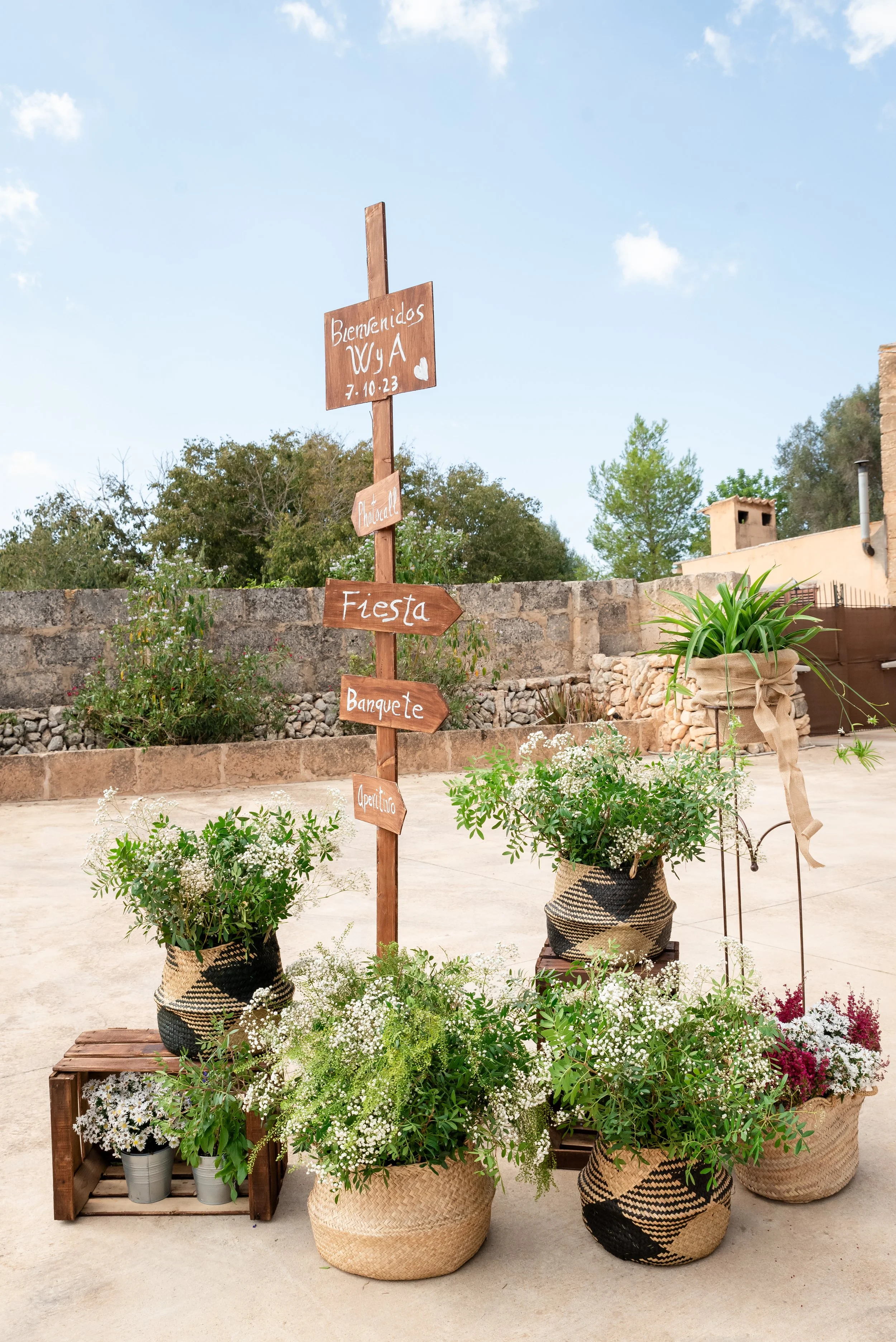 Decoración de boda al aire libre con un letrero de madera indicando "Bienvenidos", "Fiesta", "Banquete" y "Aperitivo". Hay cestas tejidas con flores y plantas verdes alrededor.