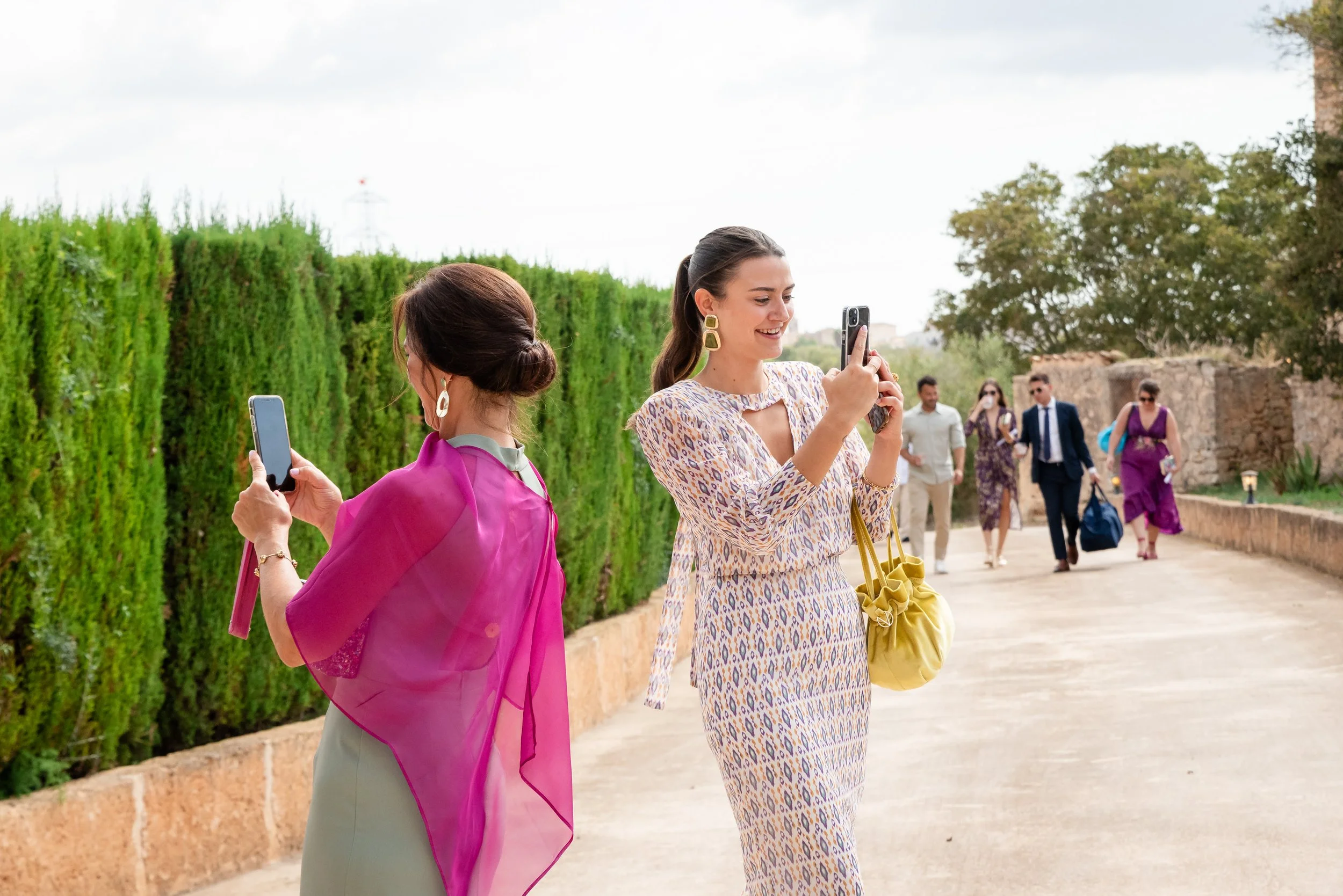 Personas bien vestidas tomando fotos con sus teléfonos en un camino al aire libre, rodeadas de arbustos y árboles; otras personas caminando al fondo.