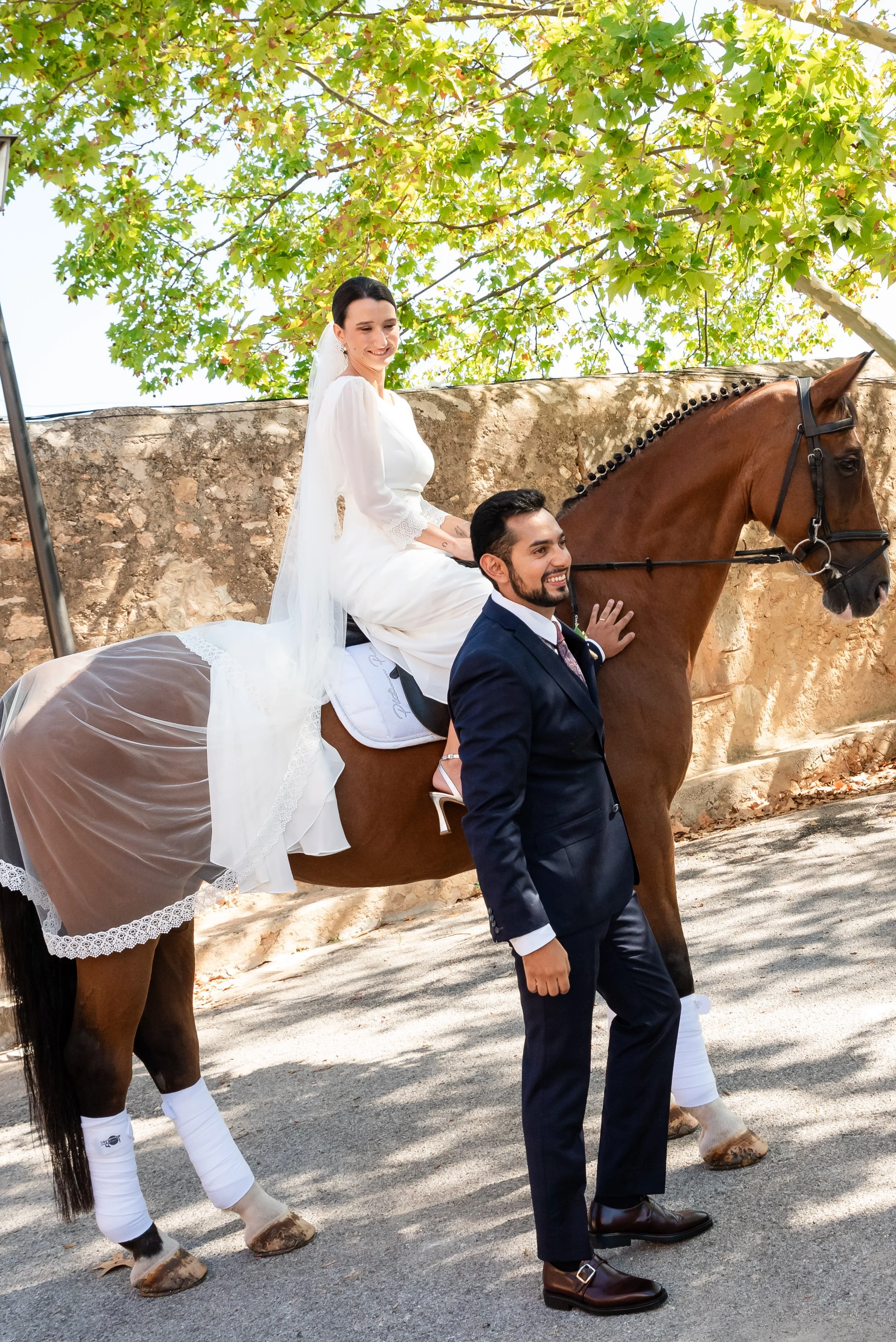 Pareja de recién casados, la novia vestida de blanco montando un caballo, y el novio al lado, ambos sonrientes, en un entorno al aire libre con árboles de fondo.