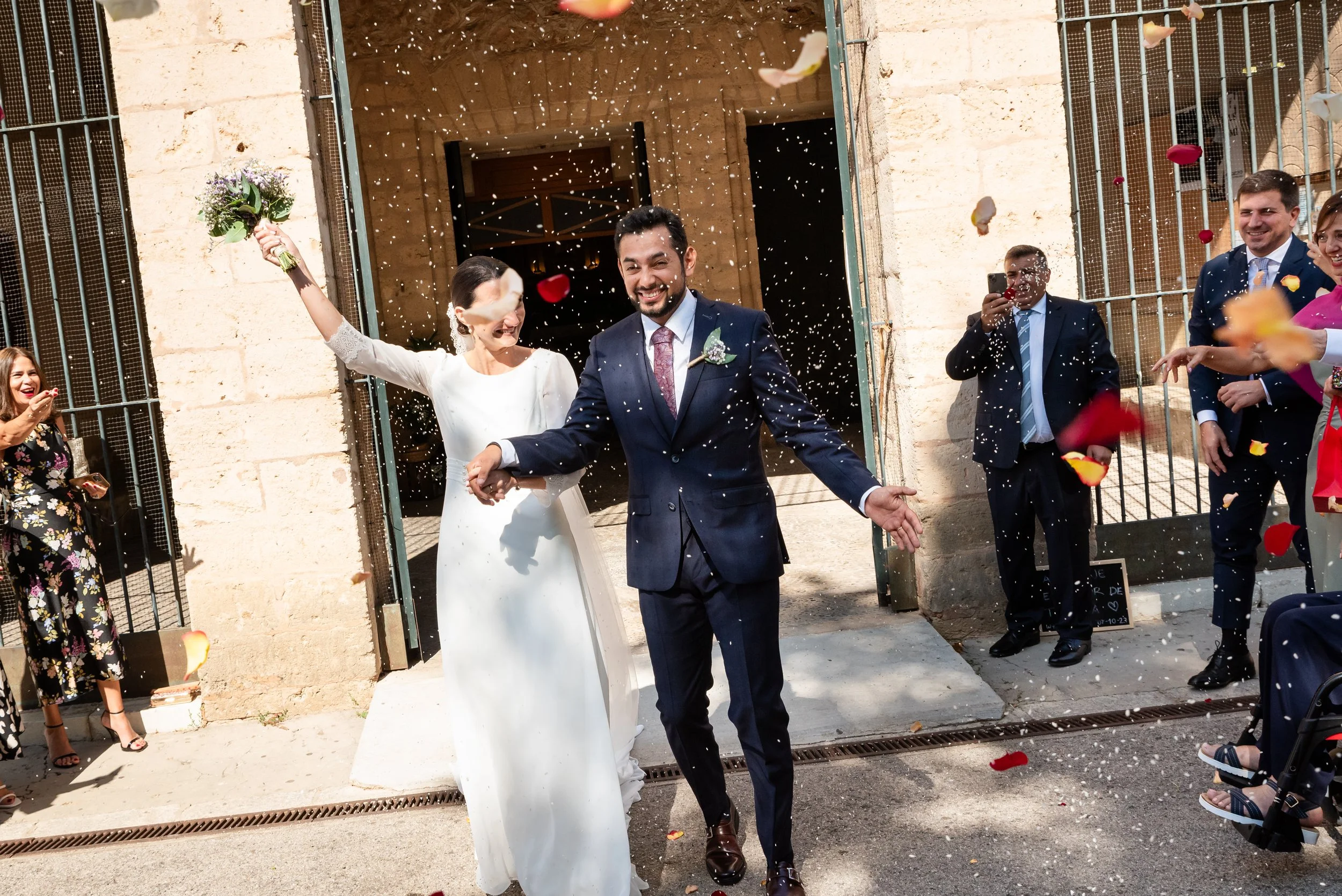 Pareja recién casada saliendo de una iglesia, rodeada de invitados lanzando pétalos y arroz.