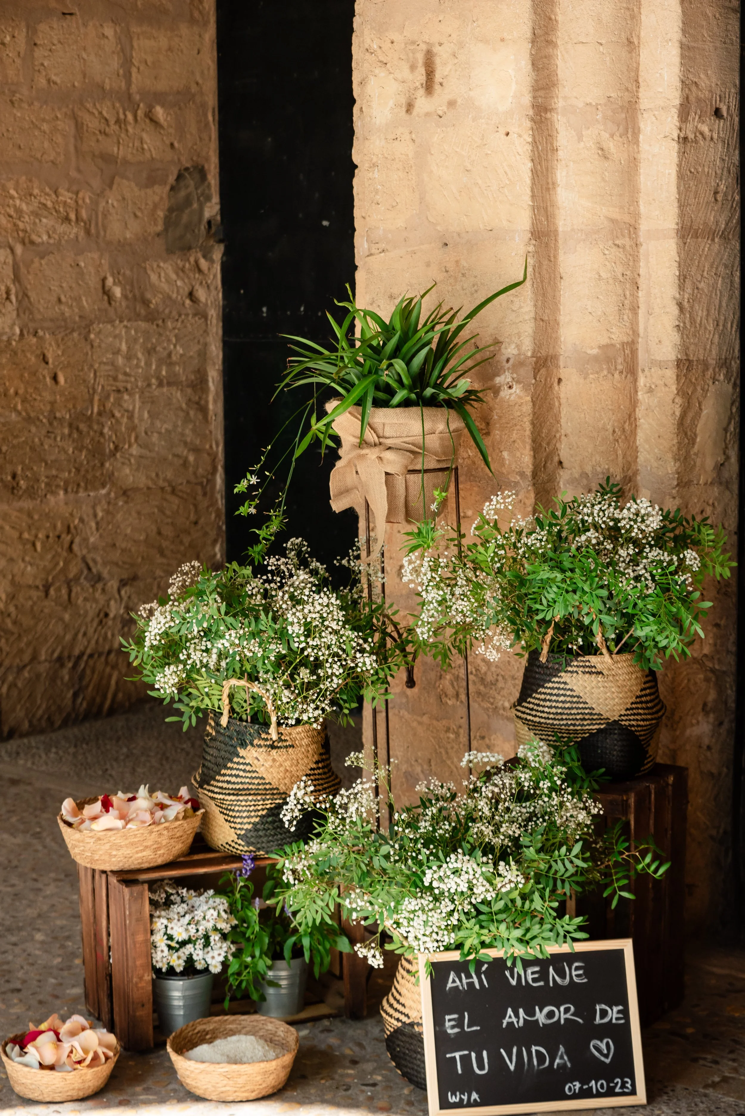 Arreglo de plantas en cestas de mimbre contra una pared de piedra, con un cartel que dice "AHÍ VIENE EL AMOR DE TU VIDA". Hay cestas con pétalos y jarrones metálicos con flores pequeñas.