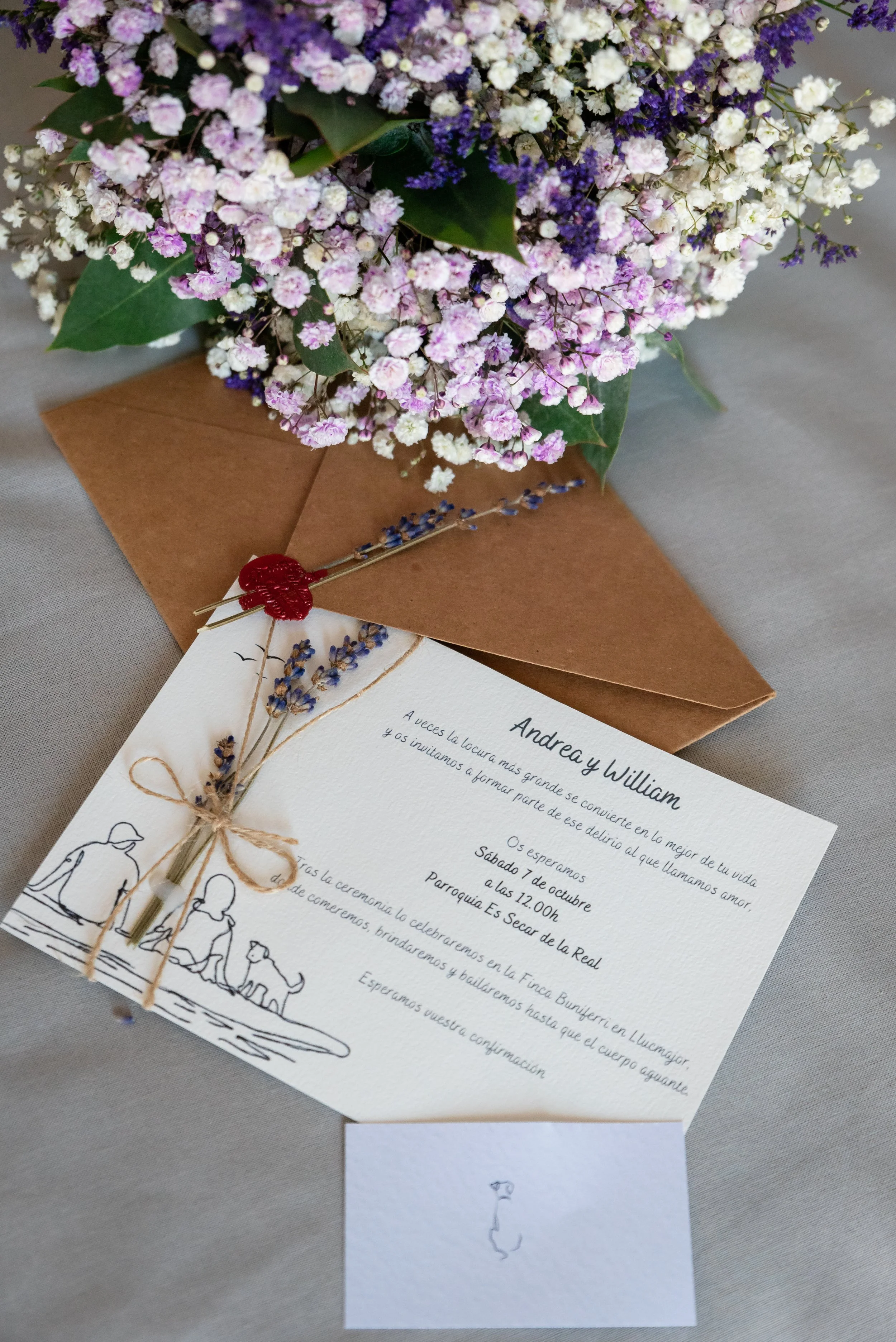 Ramo de flores blancas y moradas encima de un sobre marrón con una invitación de boda. La invitación tiene un diseño de un dibujo de una pareja con un perro y un adorno de lavanda atado con una cuerda. Hay una tarjeta pequeña con un dibujo al lado.