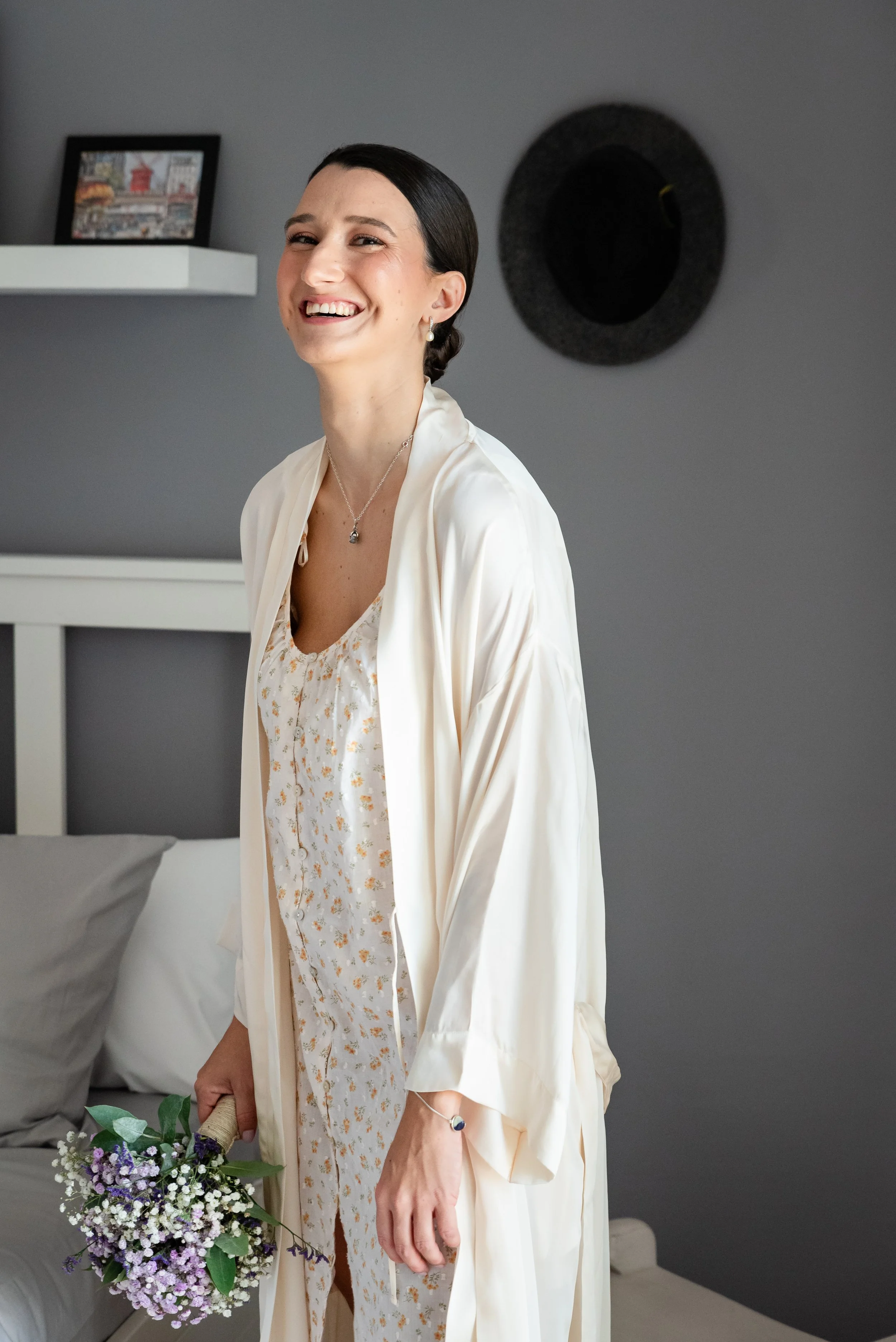 Mujer sonriente con bata de flores blancas, sosteniendo un ramo de flores púrpuras, en un dormitorio con paredes grises.