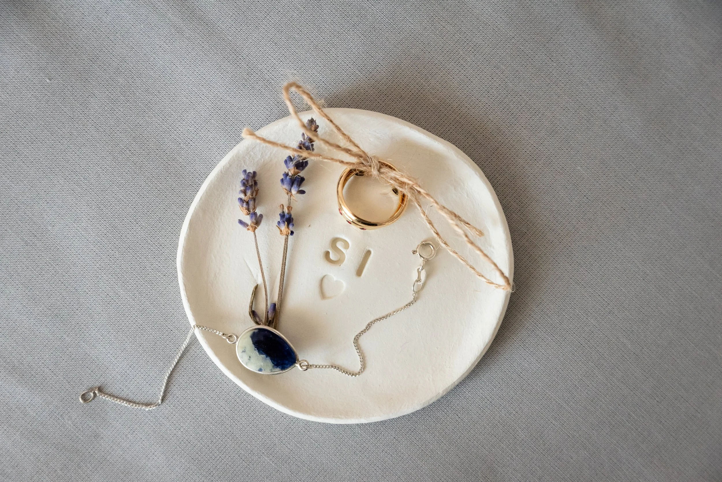 Plato de cerámica con ramo de lavanda, anillo dorado atado con cuerda y dije de piedra azul con cadena, sobre fondo gris.