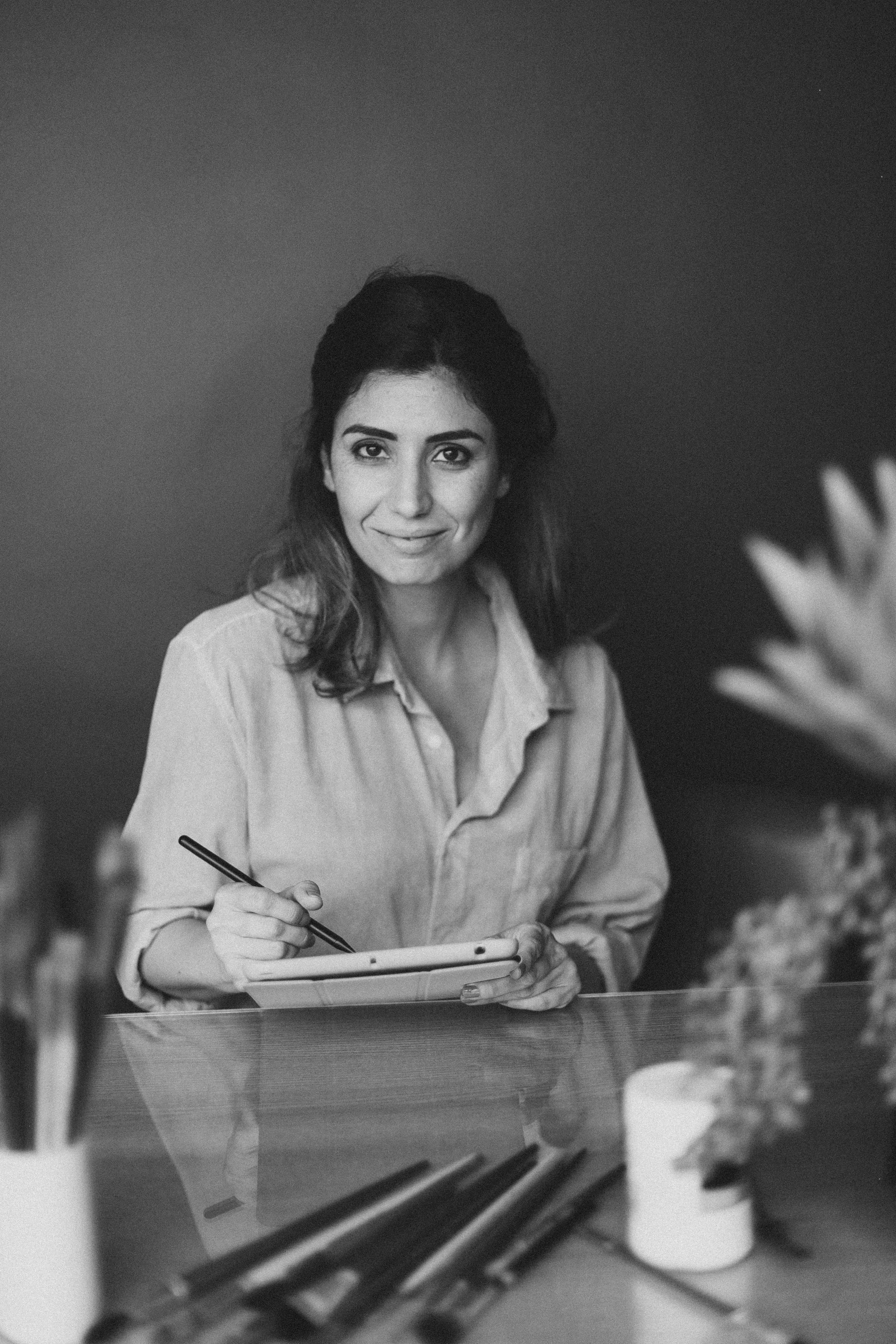 Mulher sorrindo segurando uma caneta e um tablet, com pincéis em cima de uma mesa. Ambiente de trabalho artístico, imagem em preto e branco.
