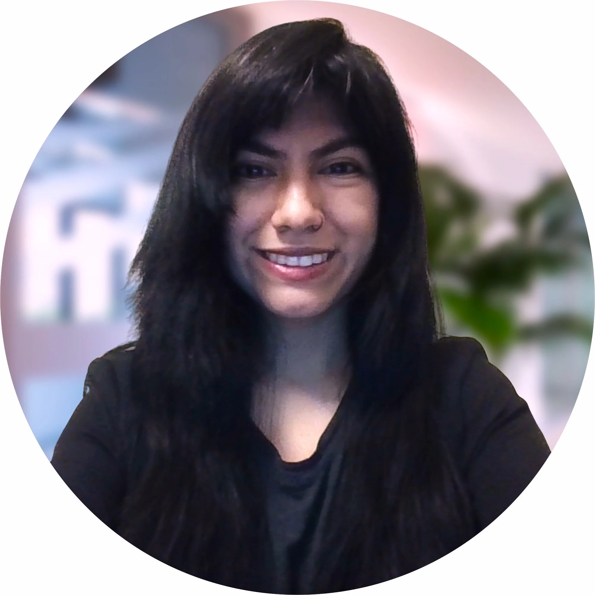 A young woman with long black hair, smiling in an indoor setting, with plants in the background.