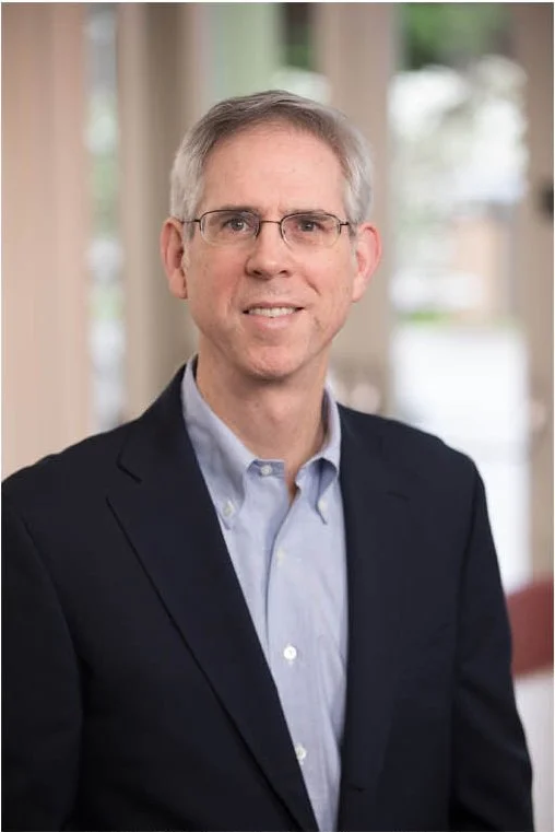 A middle-aged man with gray hair, glasses, and a friendly expression, dressed in a dark suit jacket and light blue collared shirt, standing indoors in front of windows with blurred background.