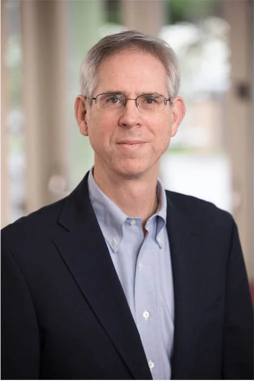 Portrait of a middle-aged man with short gray hair, glasses, wearing a dark blazer over a light blue dress shirt, standing outdoors with a blurred background.