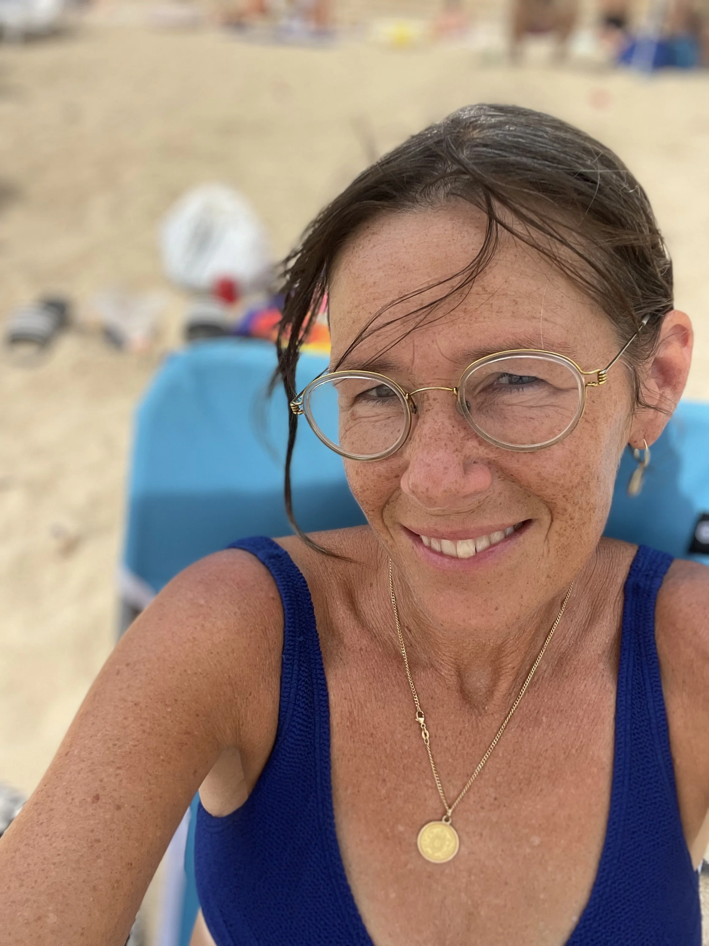 Kvinna med glasögon och halsband på stranden med sand och strandutrustning i bakgrunden.