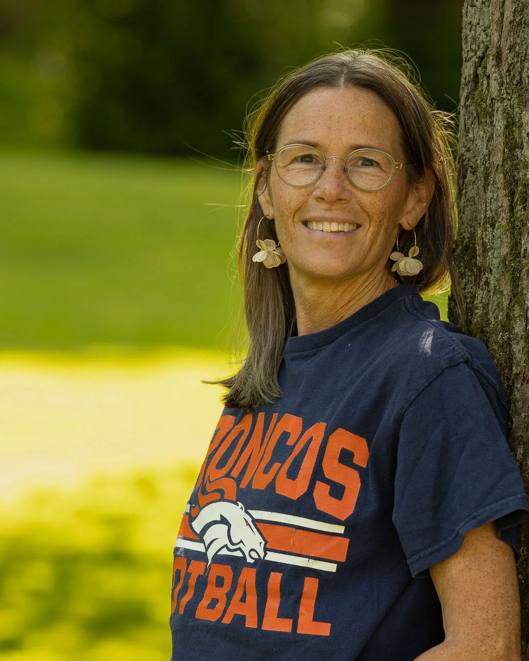 Kvinna som lutar sig mot ett träd, utomhus, med grön bakgrund, bär glasögon, stora örhängen med blomformat motiv och en Denver Broncos Football T-shirt.