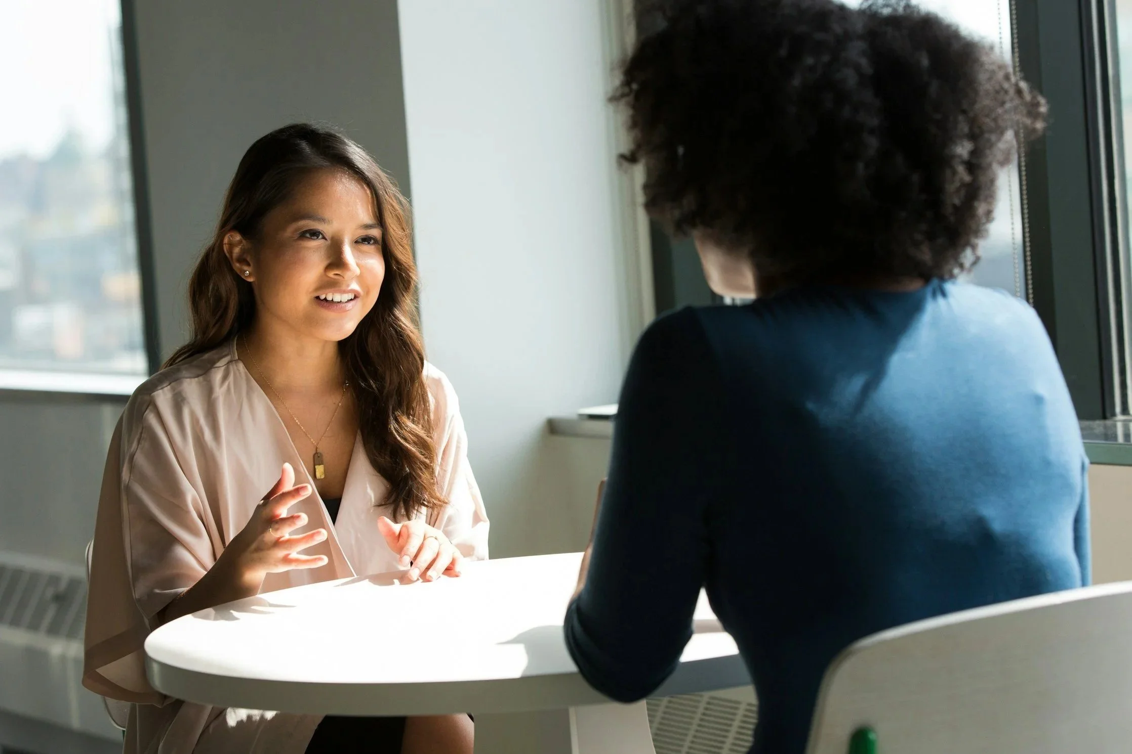 Two women having a conversation at a table in a bright room with large windows.