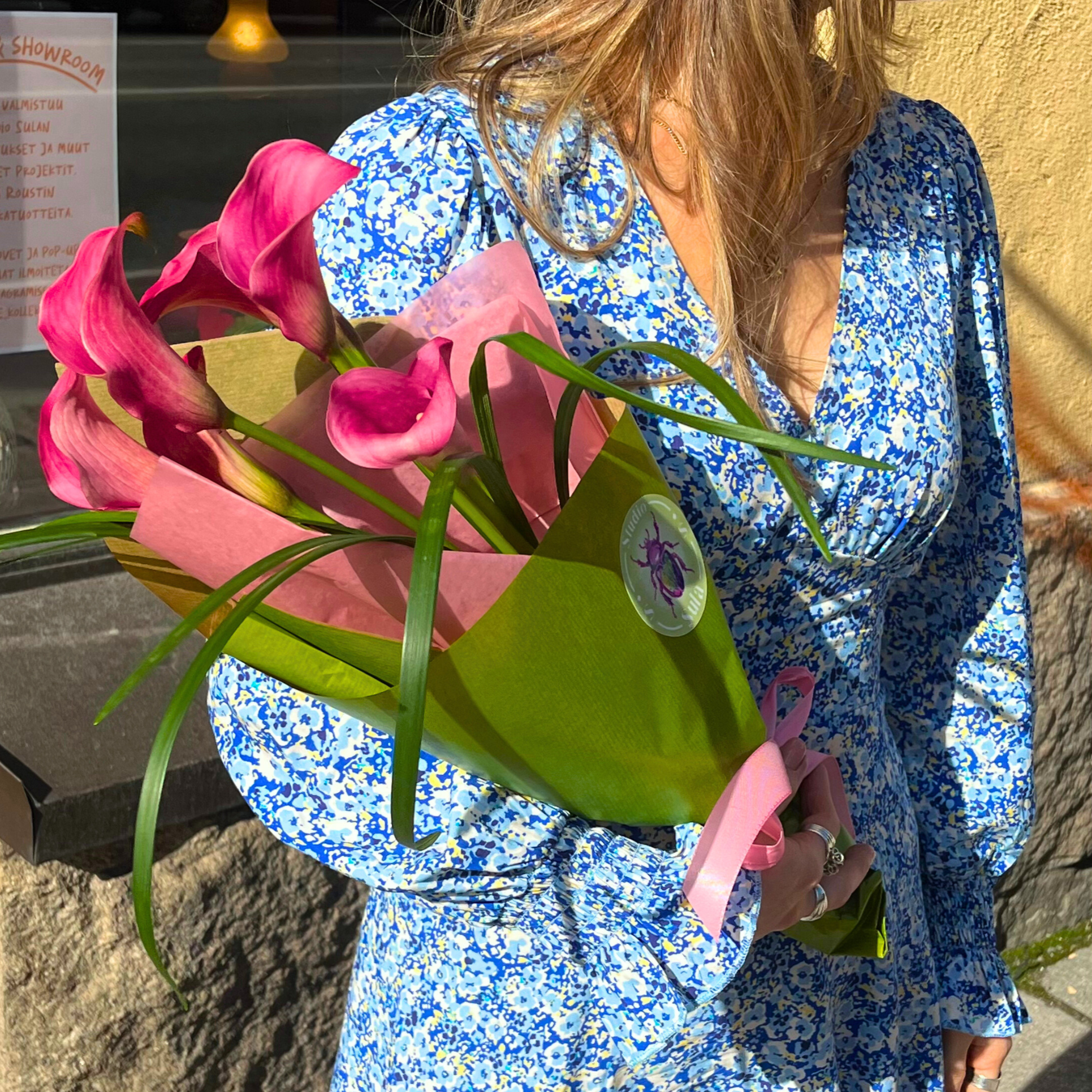 Nainen, joka pitää kukkakimppua, istuu ulkona. Hänellä on päällään sininen, valkoisella ruudukkokuviolla oleva mekko ja hänellä on värillisiä rihkamakoruja.