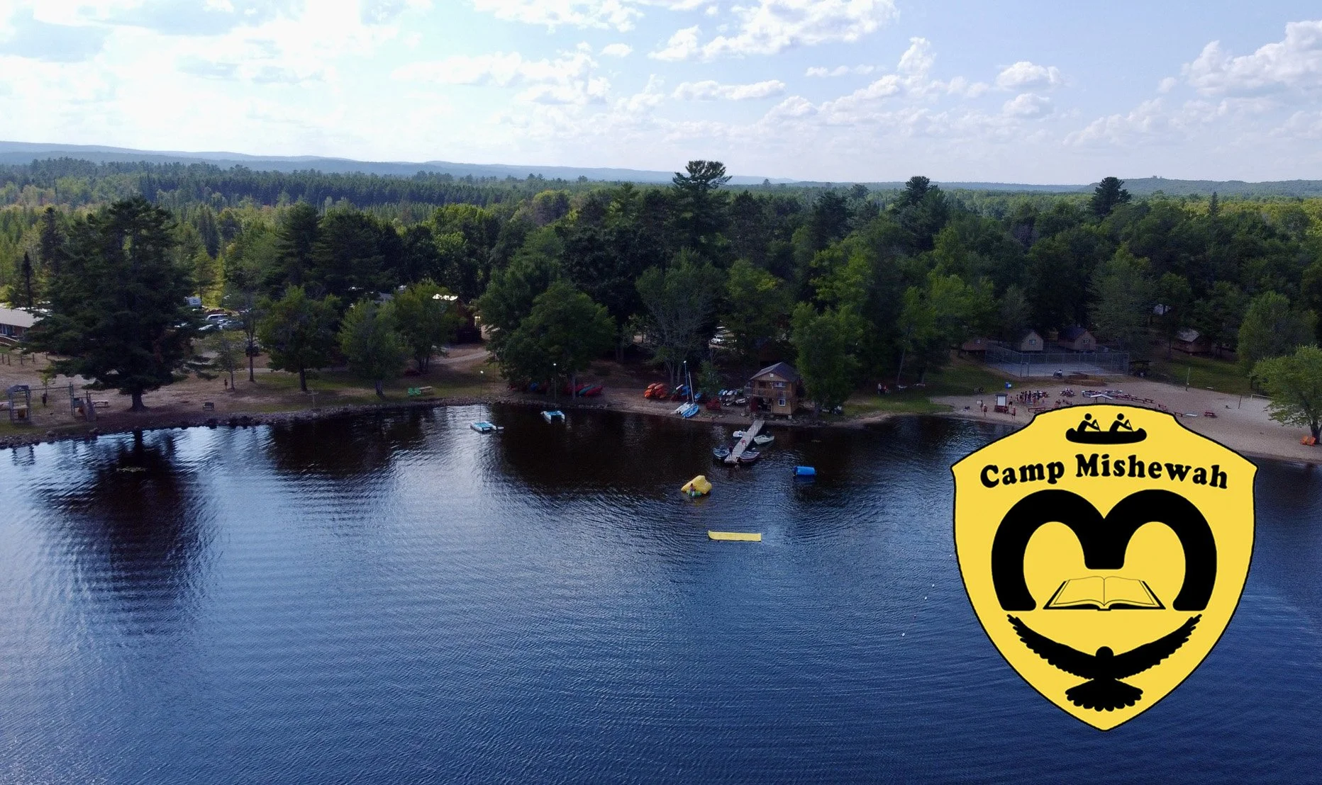 Aerial view of children and adults forming the words "CAMP" on sandy beach beside a body of water, surrounded by boats and kayaks, with trees in the background and a Camp Mishewah logo in the corner.