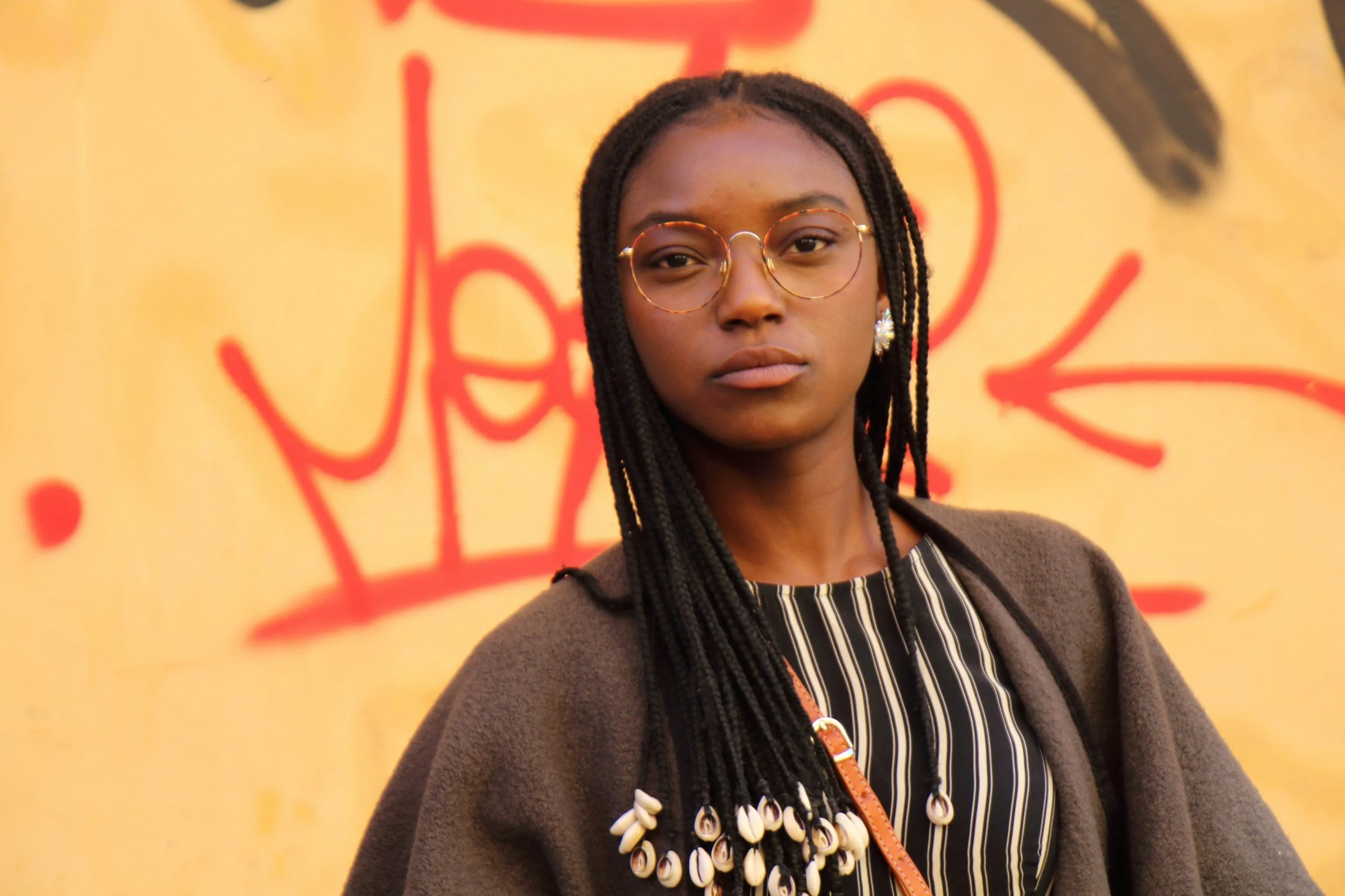 Person with braided hair and glasses standing in front of a graffiti wall.