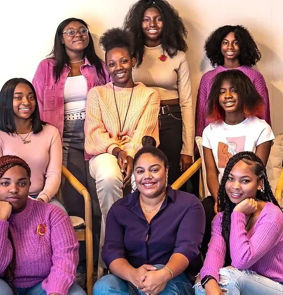 Group of nine smiling people, mostly women, sitting and standing in casual clothing, with diverse hairstyles and colorful outfits, posing together indoors.