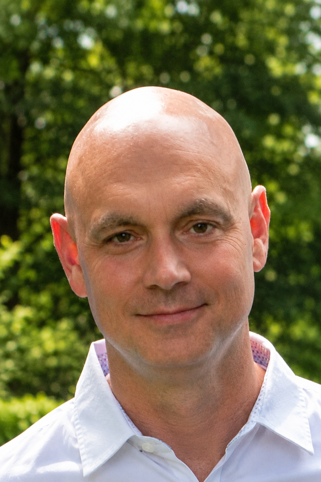Bald man smiling outdoors with green foliage background