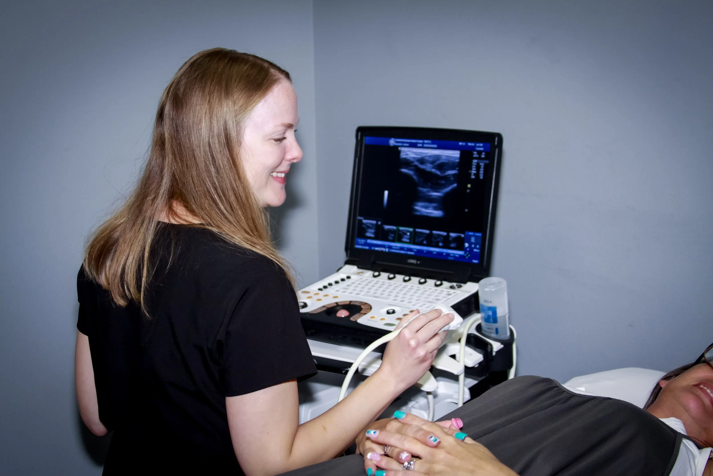 A woman with long red hair wearing a black shirt is smiling while operating an ultrasound probe on a person lying down. A computer screen displaying an ultrasound image is next to her.