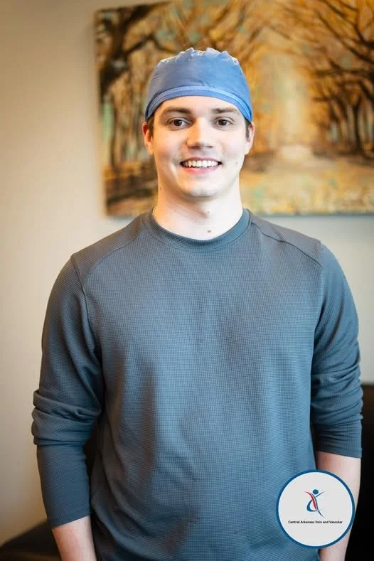 Young man in gray sweatshirt and blue surgical cap smiling indoors, with a landscape painting on the wall behind him.