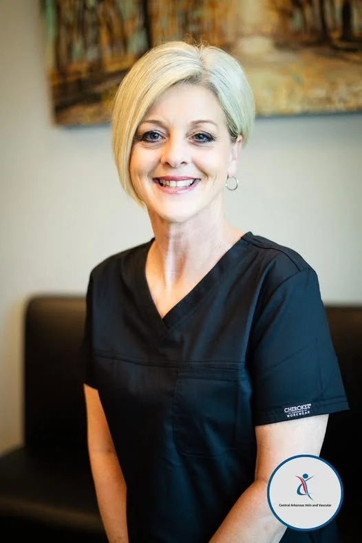 A smiling woman with short blonde hair wearing black medical scrubs, seated in a room with a colorful abstract painting on the wall behind her.