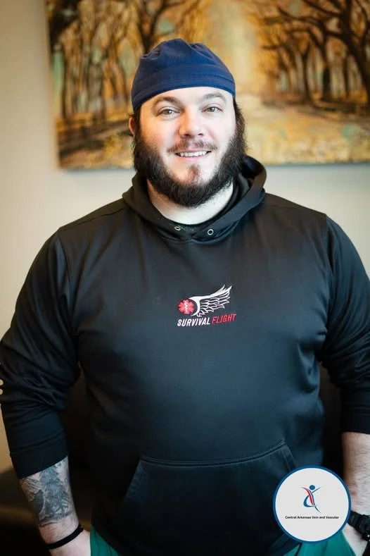 A smiling man with a beard wearing a blue beanie and black hoodie with a logo that says 'Survival Flight.' There is a colorful painting of trees and a pathway in the background and a circular sticker for the Central Arkansas Vets and Vascular on his 