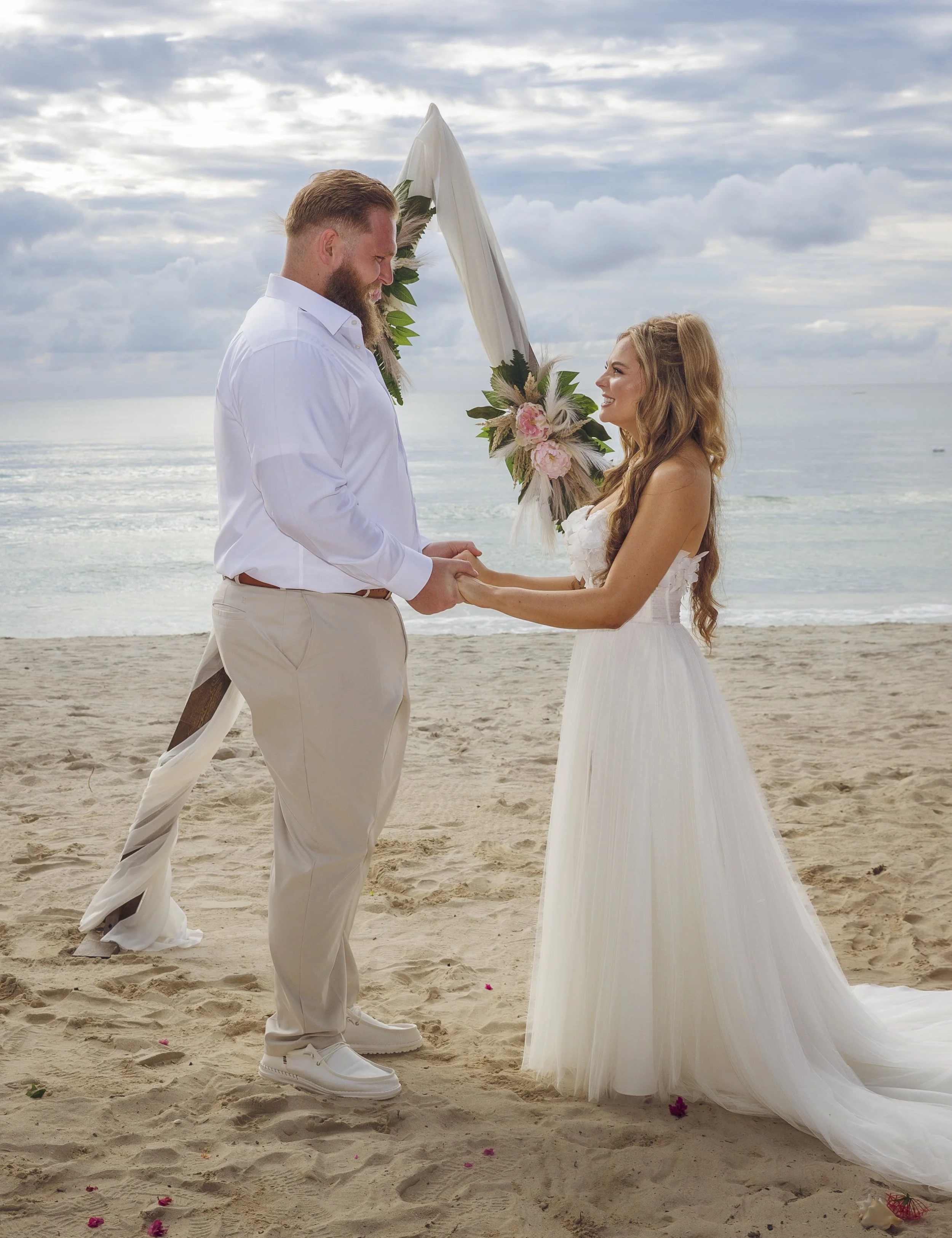 A couple exchanging vows during a beach wedding, holding hands, with a floral arch and ocean in the background.