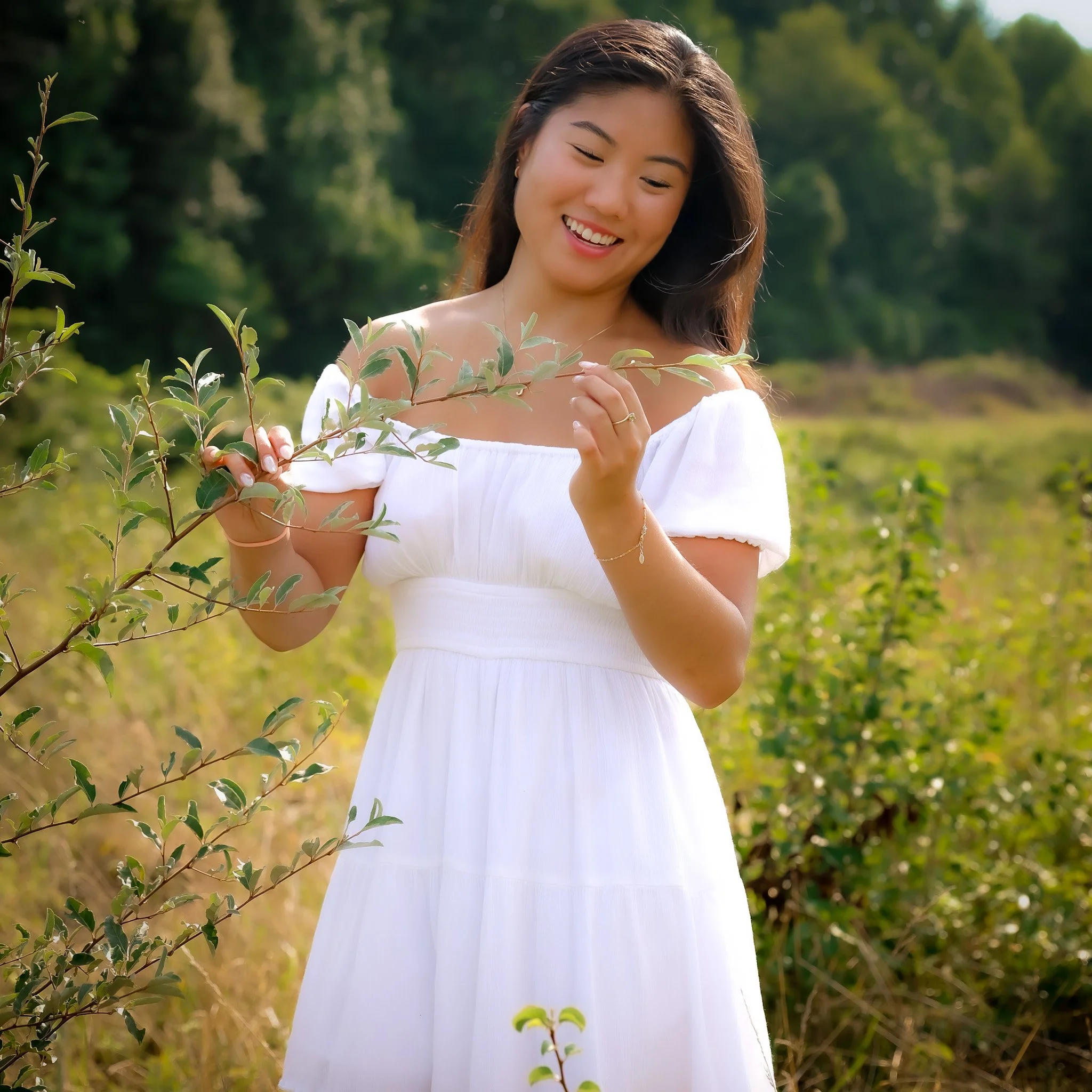 A woman in a white dress standing outdoors in a field with green bushes and trees, holding and examining a branch with leaves, smiling.
