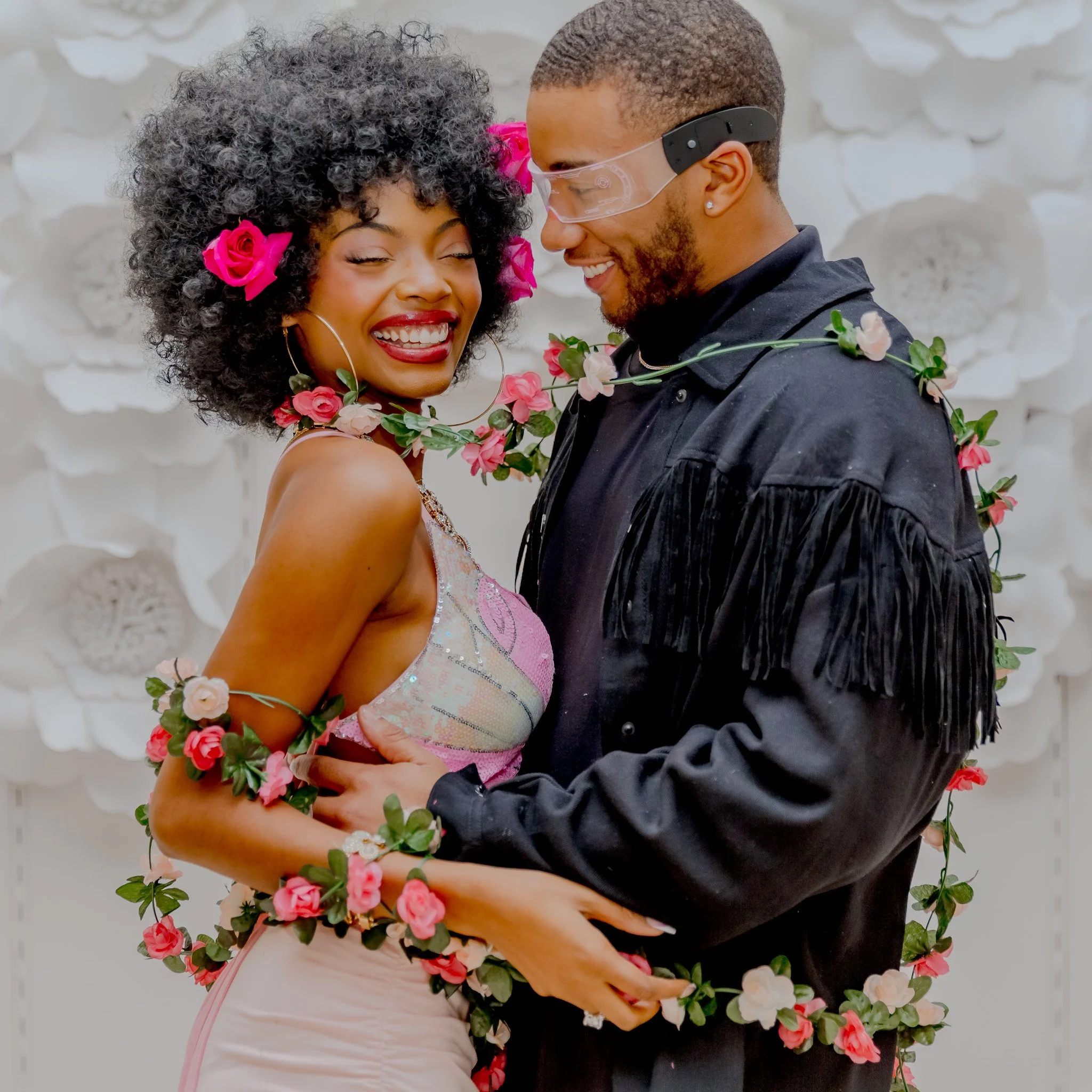 A smiling woman with curly hair decorated with pink flowers, wearing a pastel dress, embracing a man dressed in a black jacket with fringe details and glasses, both wrapped in a floral garland. The background features large white paper flowers.