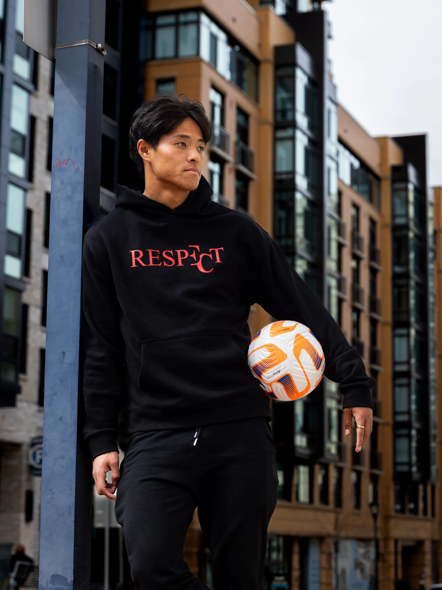A man in a black hoodie with the word "RESPECT" printed on it, holding a soccer ball, standing outdoors in an urban area with modern apartment buildings in the background.
