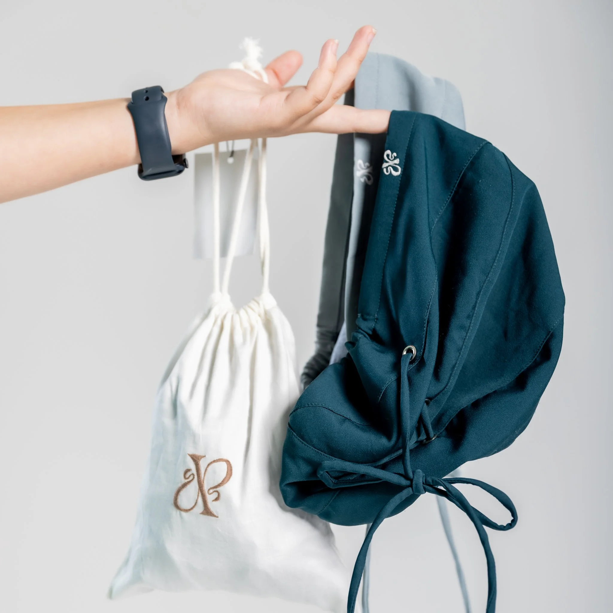 A person's hand holding a white drawstring bag and a navy blue backpack hanging on a gray coat rack against a white background.