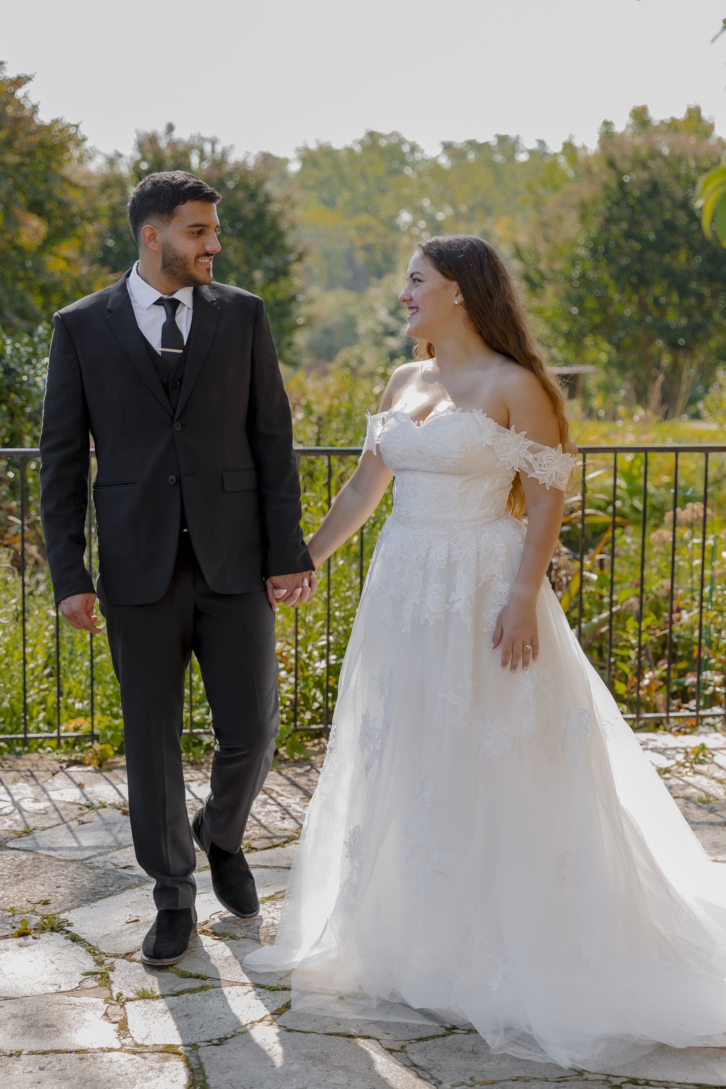 A bride and groom holding hands and smiling at each other outdoors on a sunny day.