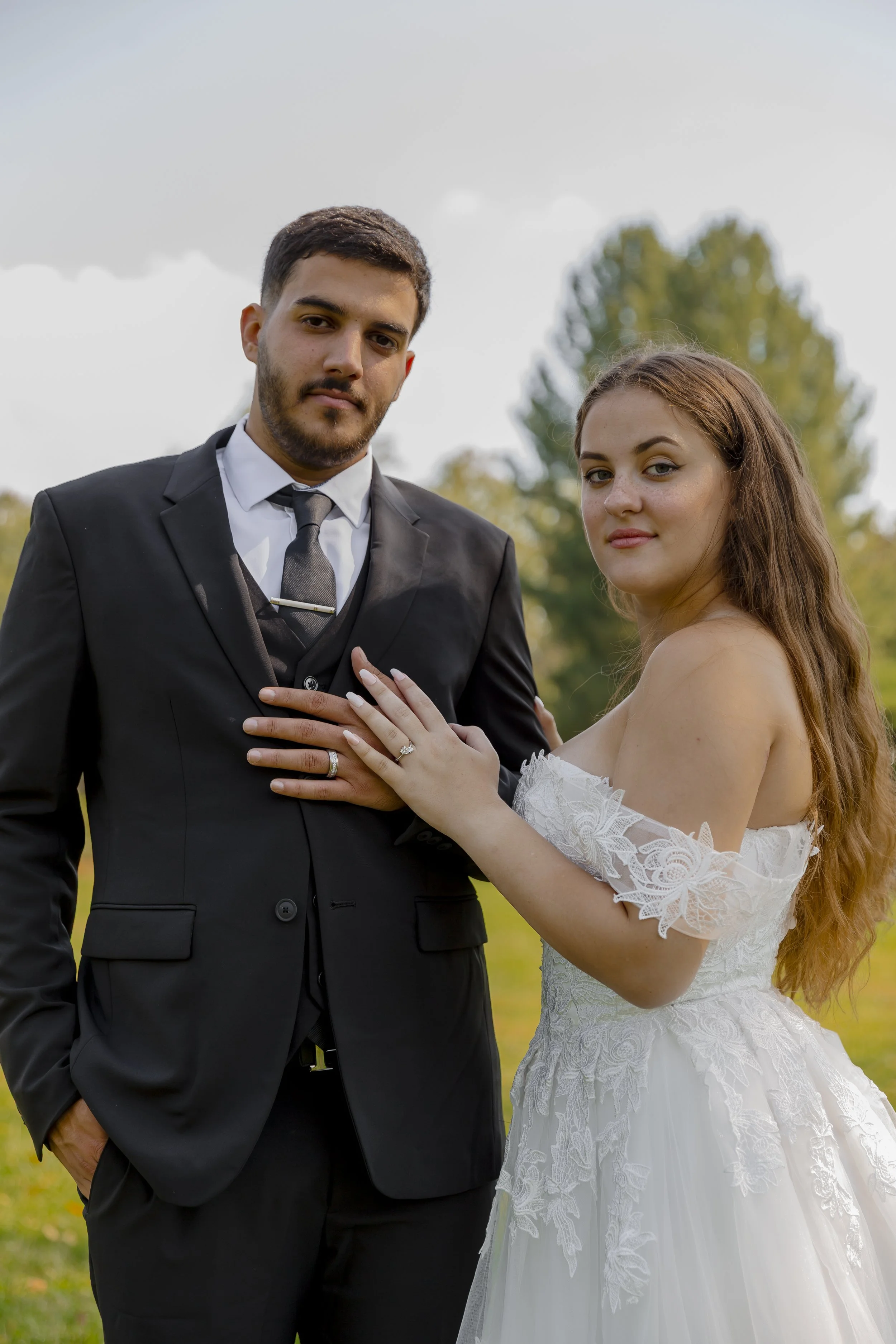 A bride and groom outdoors, the bride in a white lace wedding dress with off-shoulder sleeves and the groom in a black suit with a tie, posing for wedding photo.