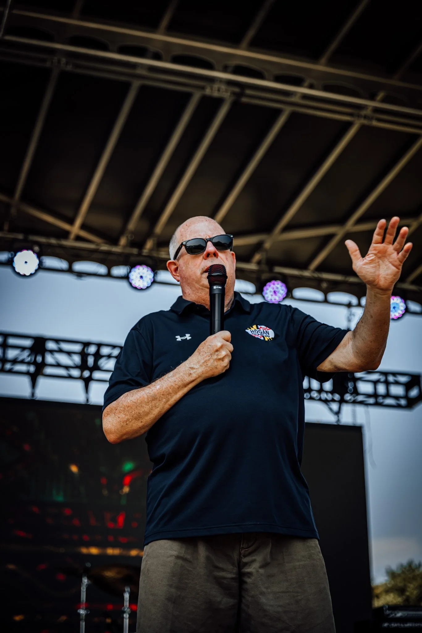 A man wearing sunglasses and a black polo shirt speaking into a microphone on an outdoor stage.