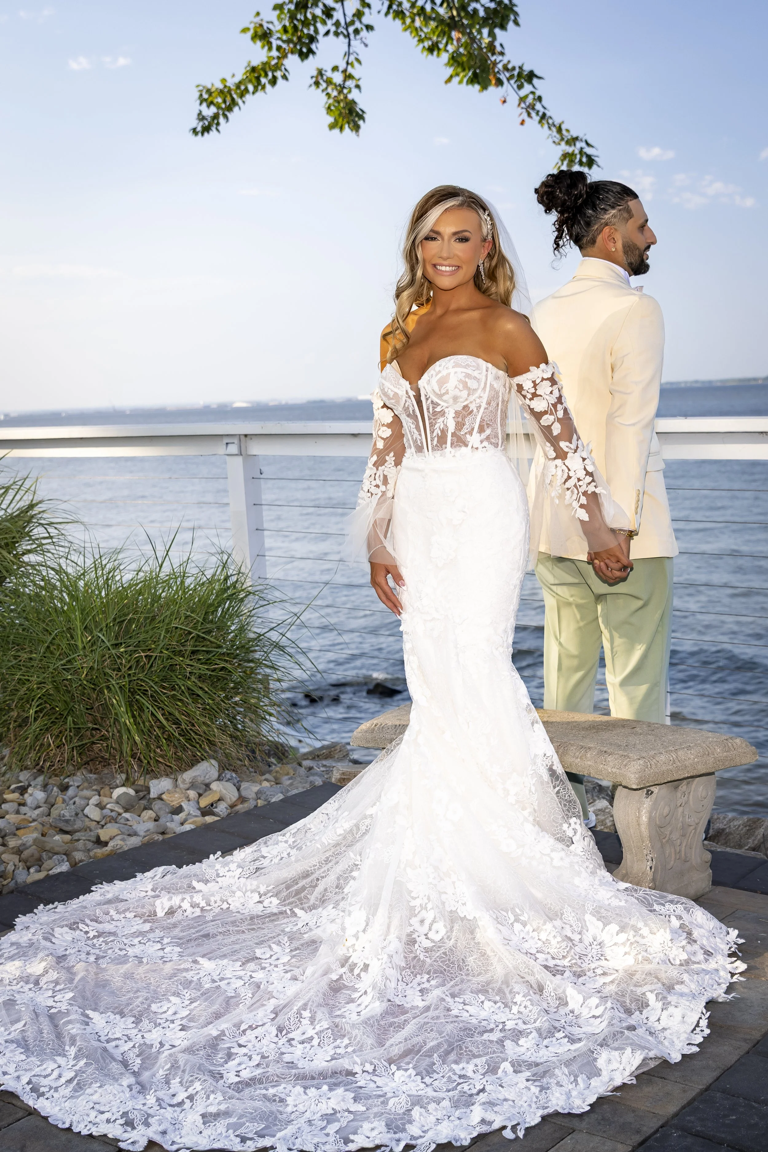 A bride in a white lace wedding gown holding hands with her groom, who is wearing a cream blazer, near a body of water with a clear sky overhead.