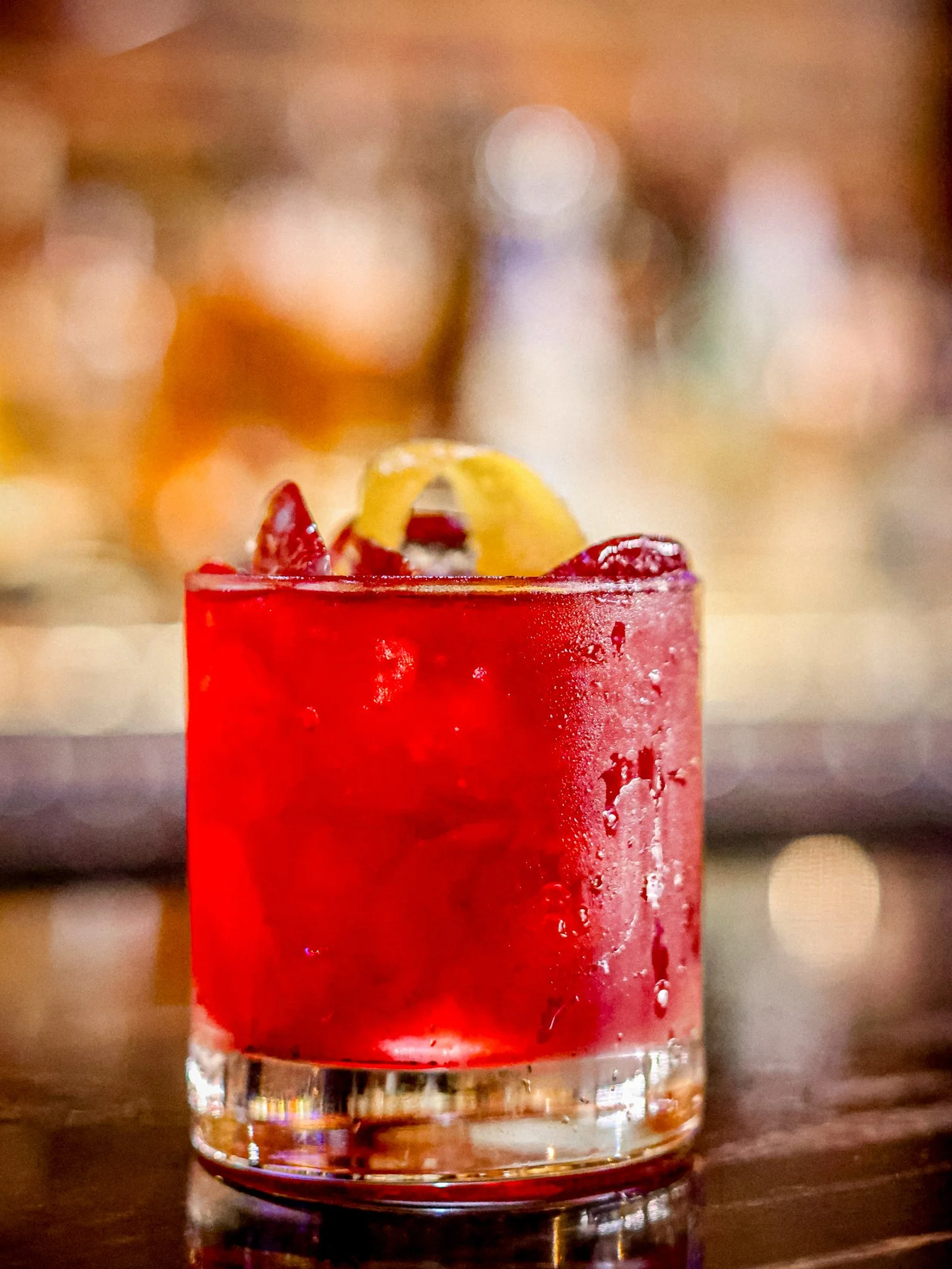 A close-up of a red cocktail in a short glass garnished with citrus peel and red fruit slices on top.