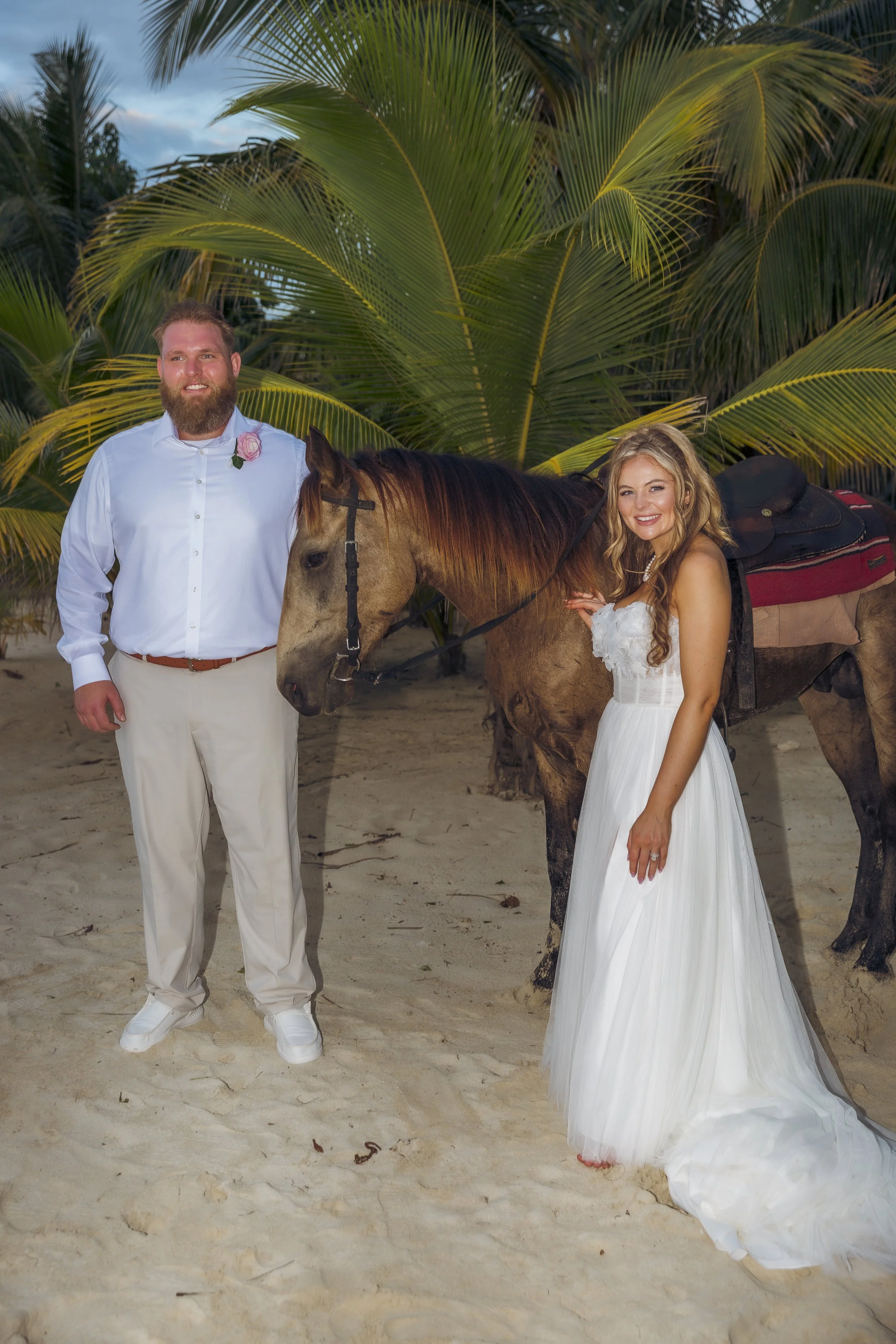 A couple dressed in wedding attire posing with a horse on a sandy beach with palm trees in the background.